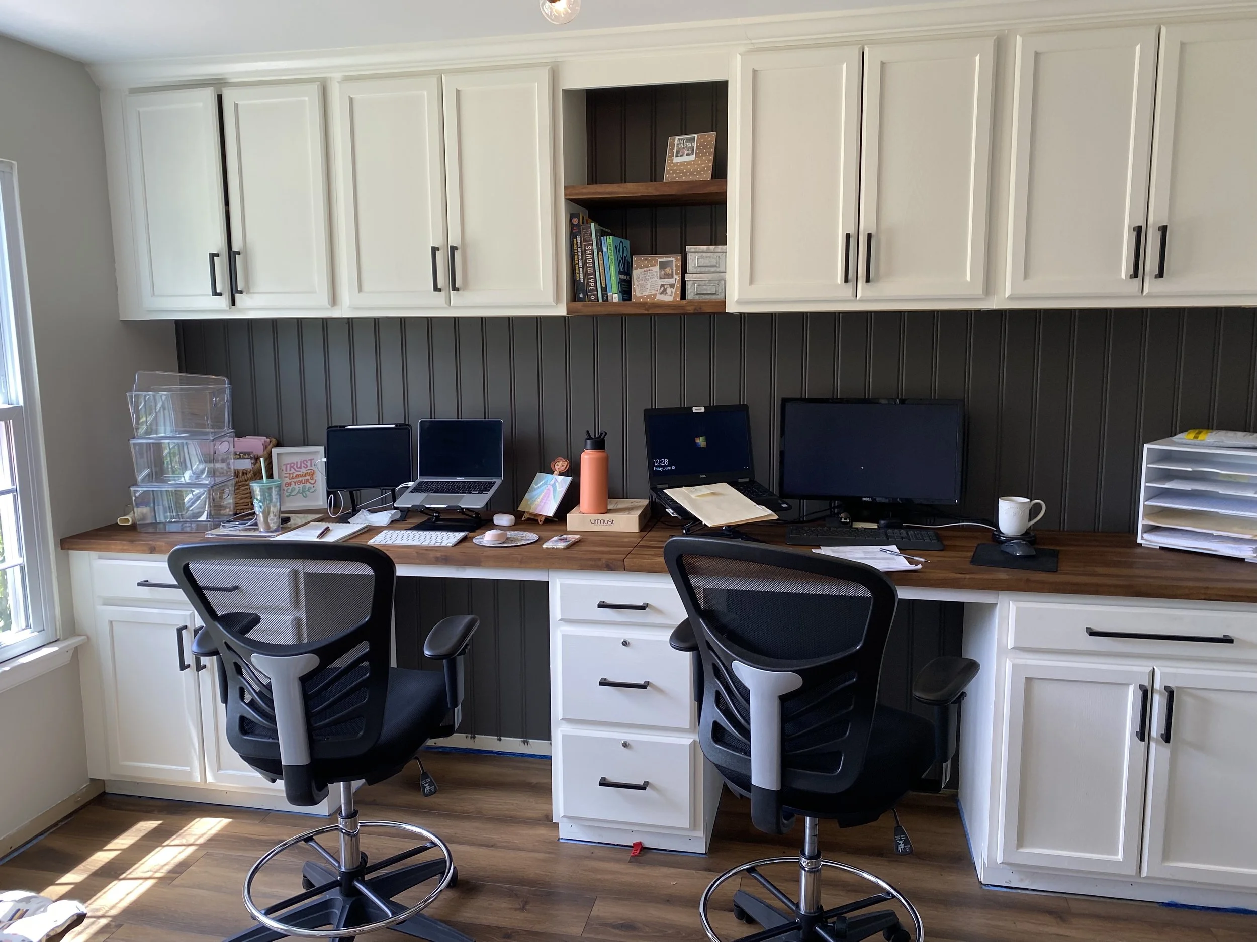 Home office with two black ergonomic chairs, a long wooden desk with two computer monitors, a laptop, office supplies, and white cabinets with black handles above the desk, and a window to the left allowing natural light.