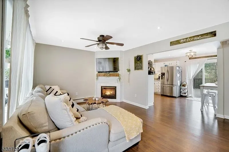 Living room with cream-colored sofa, throw pillows, a yellow blanket, hardwood floors, a wall-mounted TV above a fireplace, ceiling fan, and large windows with curtains. An open kitchen area with stainless steel refrigerator and sliding door leads outside.
