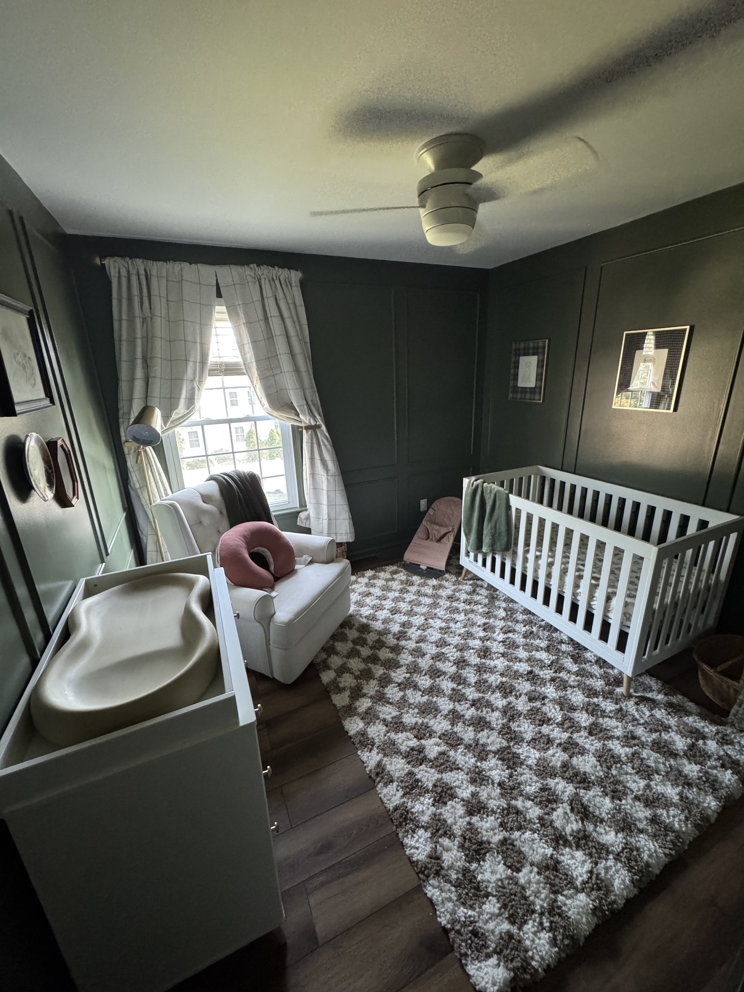 A nursery with dark green walls, a window with white plaid curtains, a white crib, a white armchair with a pink donut-shaped pillow, a pink rocking chair, a beige changing table, and a patterned rug.