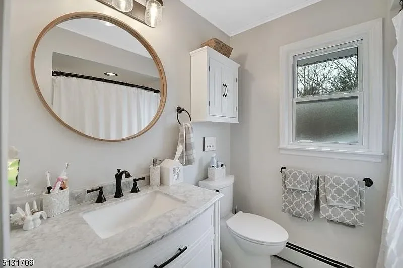 Bathroom with a white vanity, round mirror, white cabinet, toilet, window, and gray patterned towels.