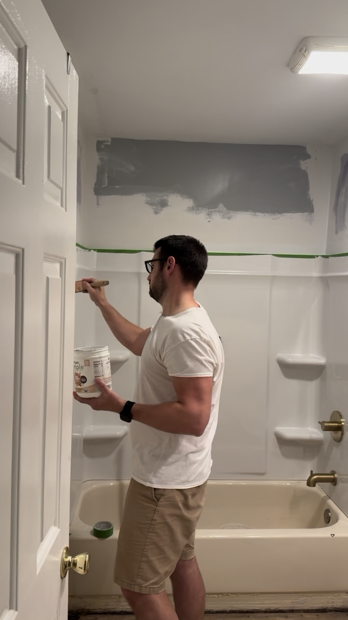A man painting a bathroom wall with primer or paint, holding a container of paint in one hand and a paintbrush in the other.