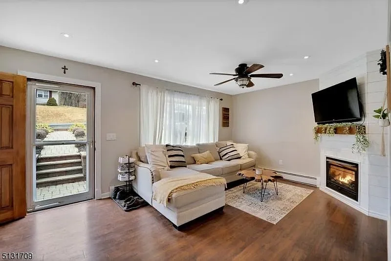 Living room with a white sectional sofa, a small wooden coffee table, a fireplace, a flat-screen TV, and a ceiling fan. Neutral-colored curtains cover the window, and there is a door leading outside.