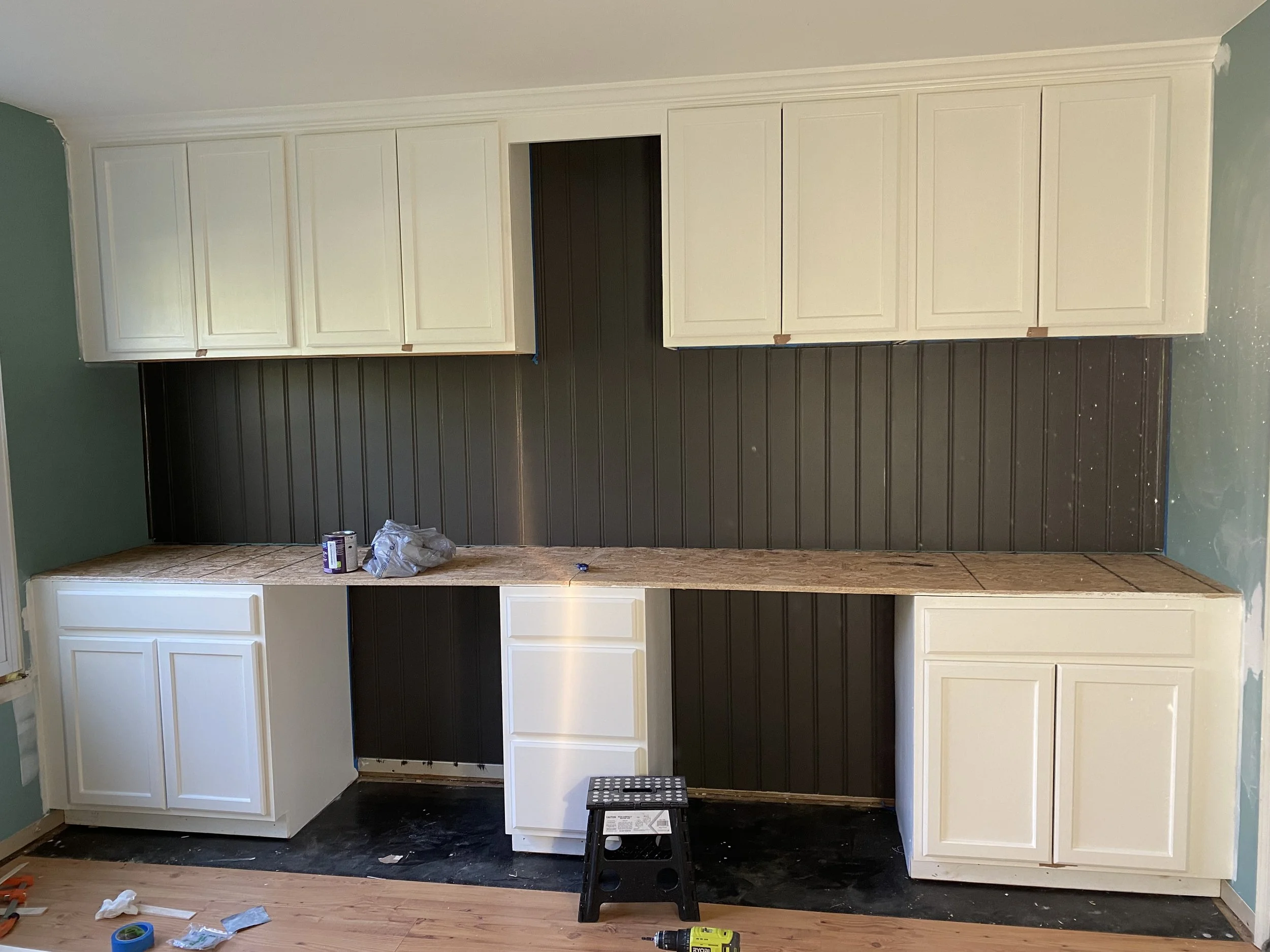 Kitchen wall with white upper cabinets and lower cabinets, black beadboard backsplash, and ongoing construction with tools and supplies on the countertop and floor.