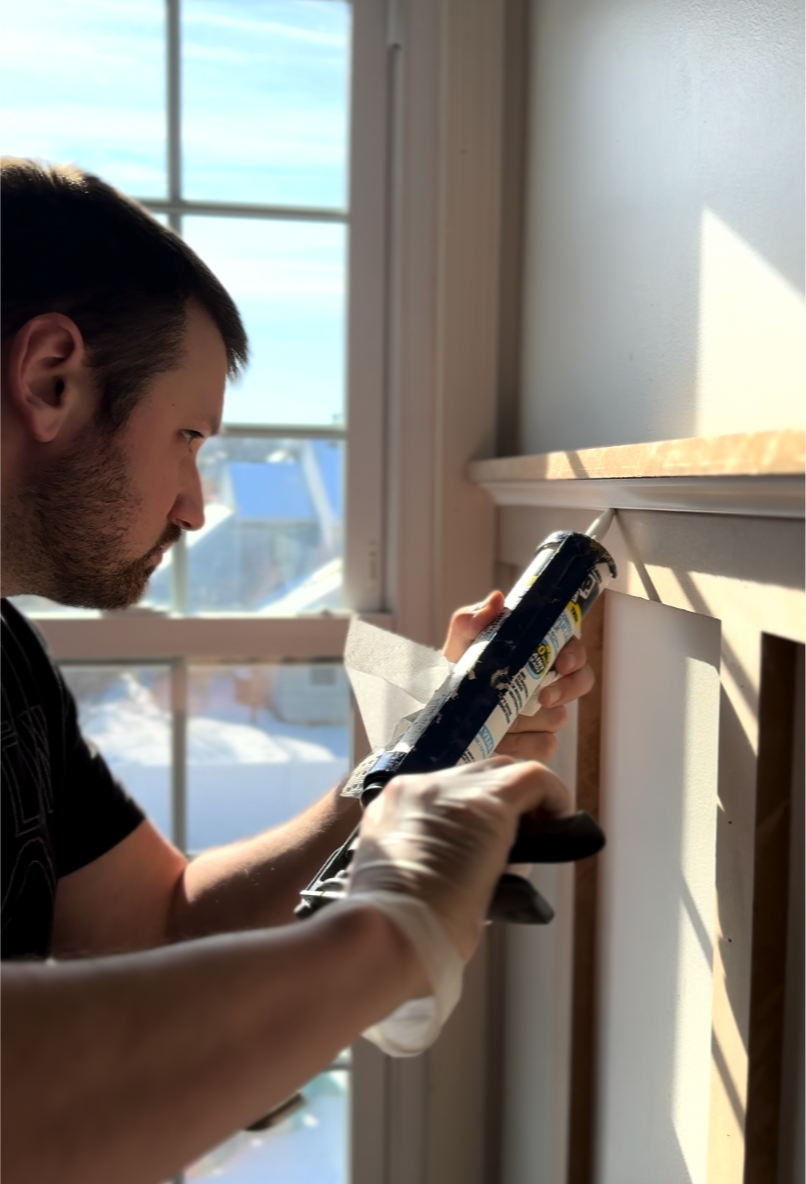 A man wearing white gloves applies adhesive to the back of a white wooden panel during a home renovation, with sunlight streaming through the window behind him.