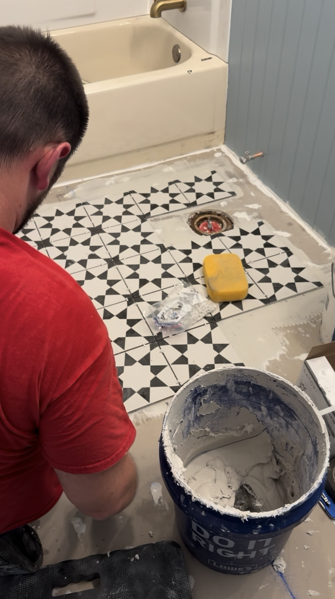 A person working on a bathroom renovation, removing old flooring and preparing to install new patterned black and white tiles. There is a bucket of white adhesive, a sponge, and tools on the floor.