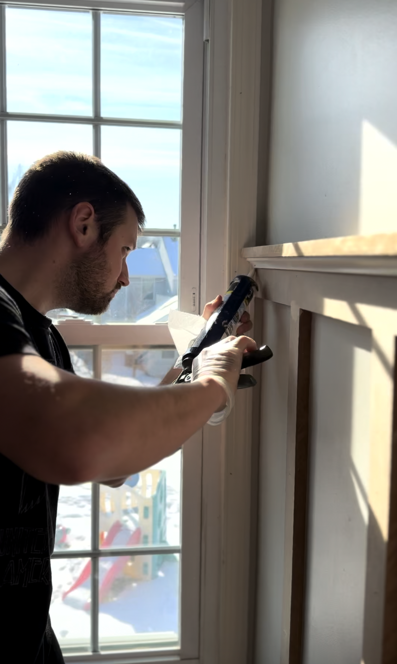 A man with a beard, wearing a black shirt and gloves, using a caulking gun to apply sealant to a wooden frame next to a window with a snowy outdoor view.