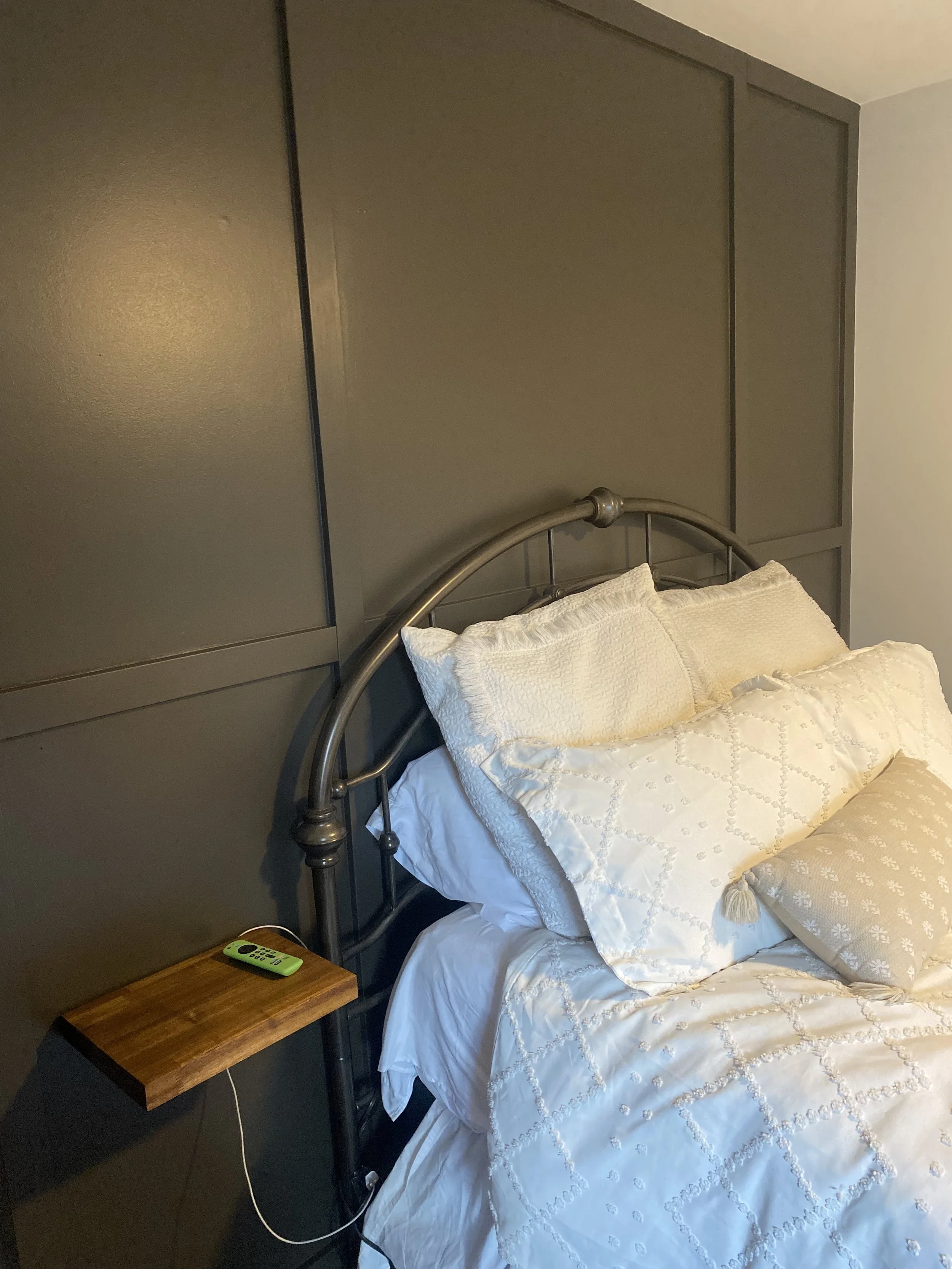 Close-up of a neatly made bed with white and cream pillows and a quilted bedspread, beside a wooden nightstand with an electronic device, against a dark gray wall with a headboard.