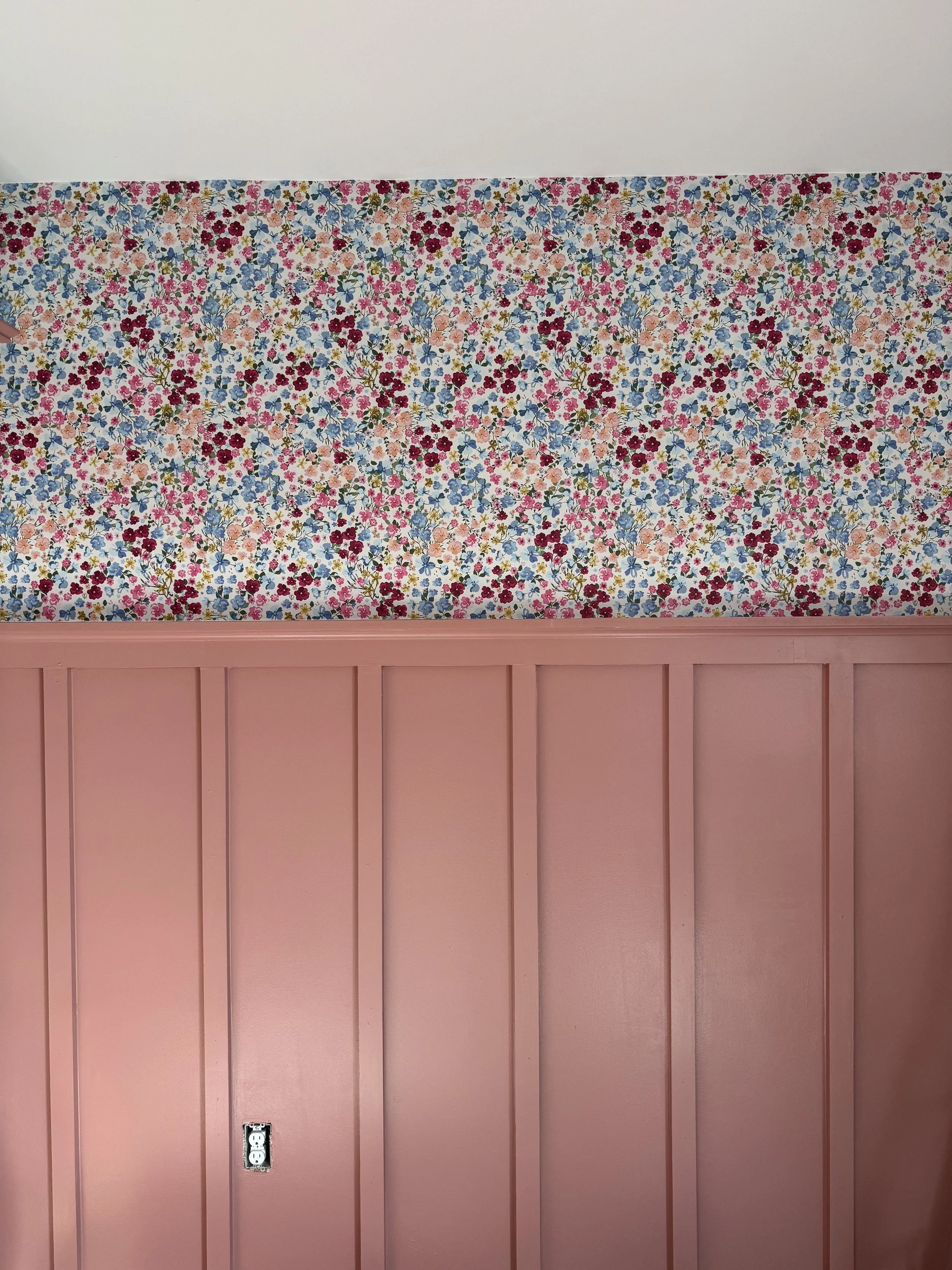A room with a white ceiling, a floral wallpaper border with colorful small flowers, and pink wall paneling with vertical grooves. There is a power outlet on the pink paneling.