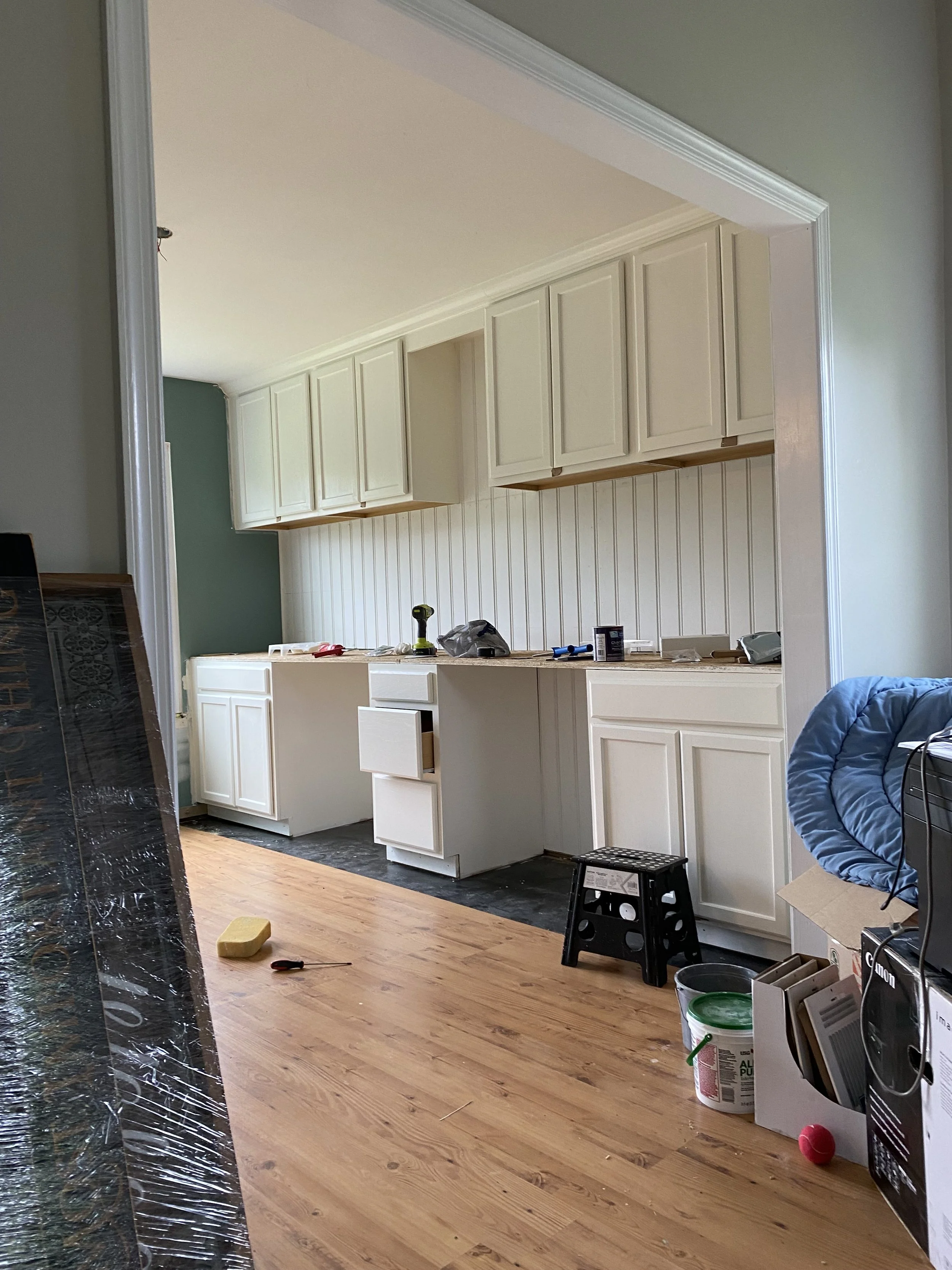 Unfinished kitchen with white cabinets, some tools and materials on the countertop, and a stool on the wooden floor.