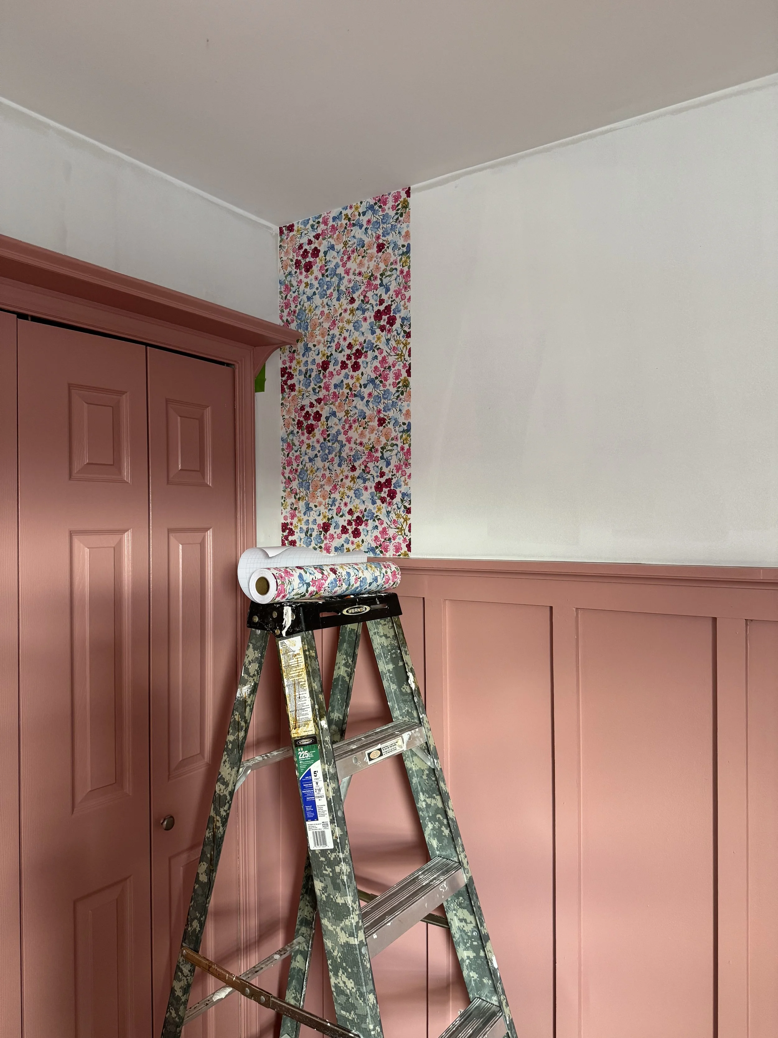 A step ladder has a roll of floral wallpaper on top, in a room with a pink wall, pink closet, and a section of floral wallpaper on the wall.