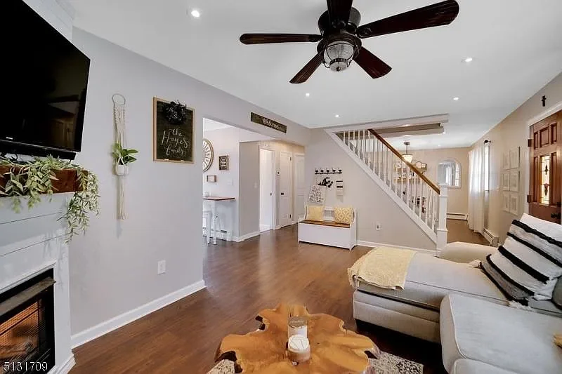 Living room with a white couch, wooden coffee table, staircase with a white railing, and a wall-mounted flat-screen TV. Decor includes a small plant, a chalkboard, and a wall-mounted clock. The room has hardwood floors, ceiling fan, and natural light
