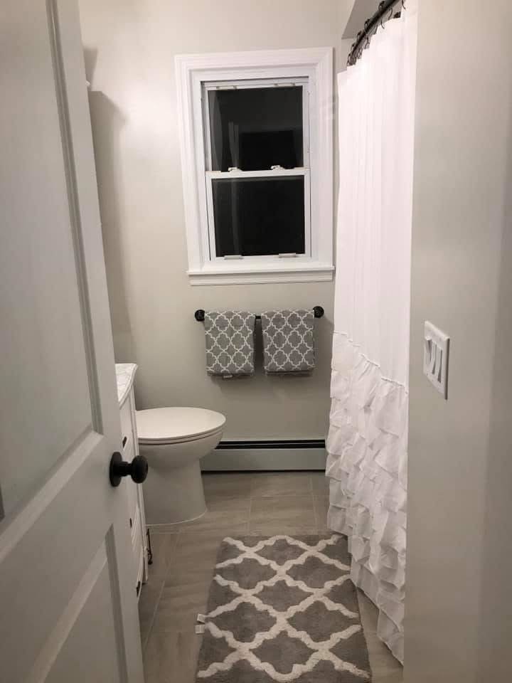 A small bathroom with a white toilet, two gray patterned towels hanging on a black towel bar, a white shower curtain with ruffles, a window above the toilet, and a gray and white patterned rug on the floor.