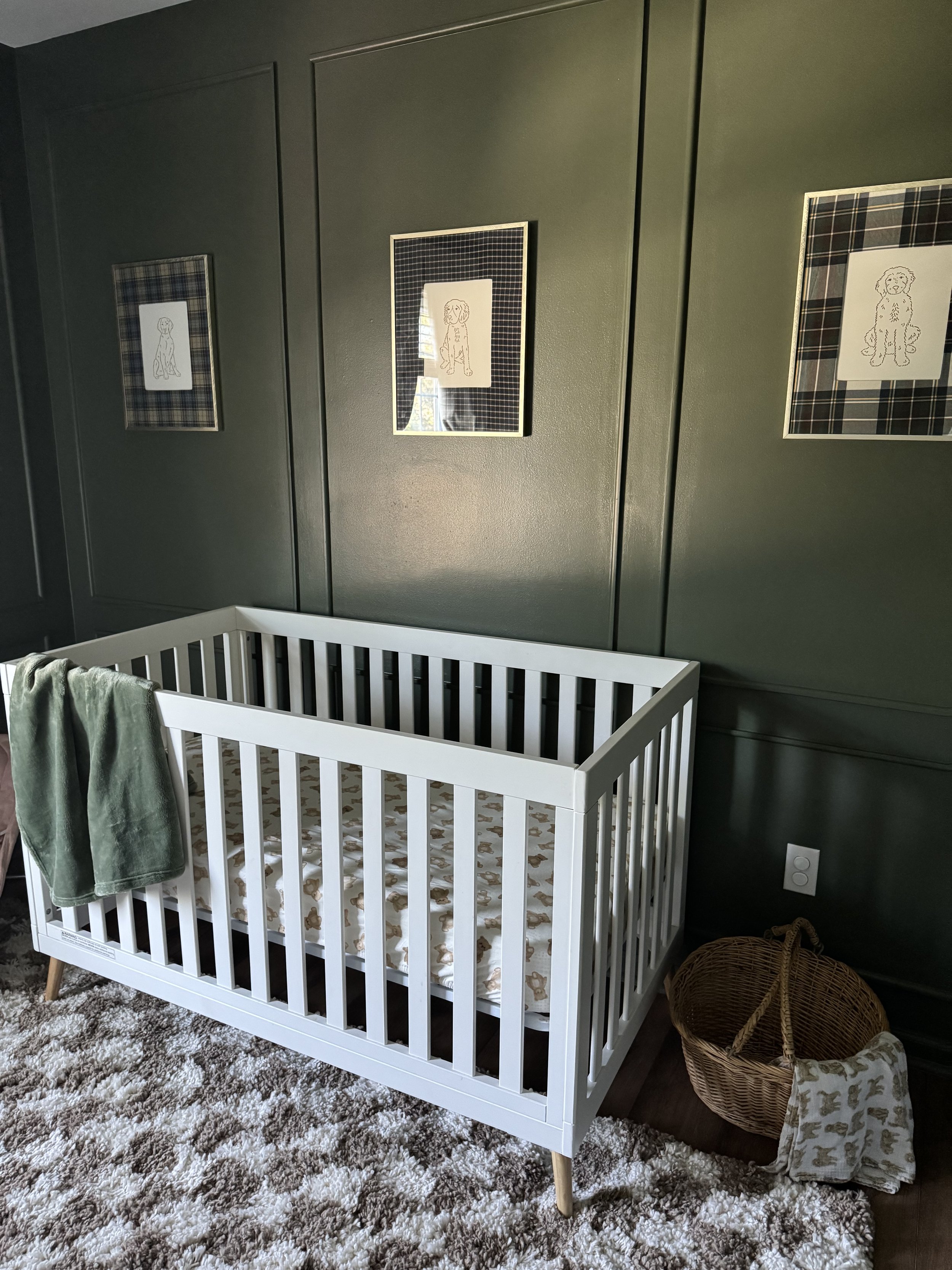 A baby crib with a white frame and a fitted sheet with a dog pattern, placed in a room with dark green wall paneling. A green towel hangs over the crib's side. There is a wicker basket on the floor next to the crib with a beige cloth inside. Three fr