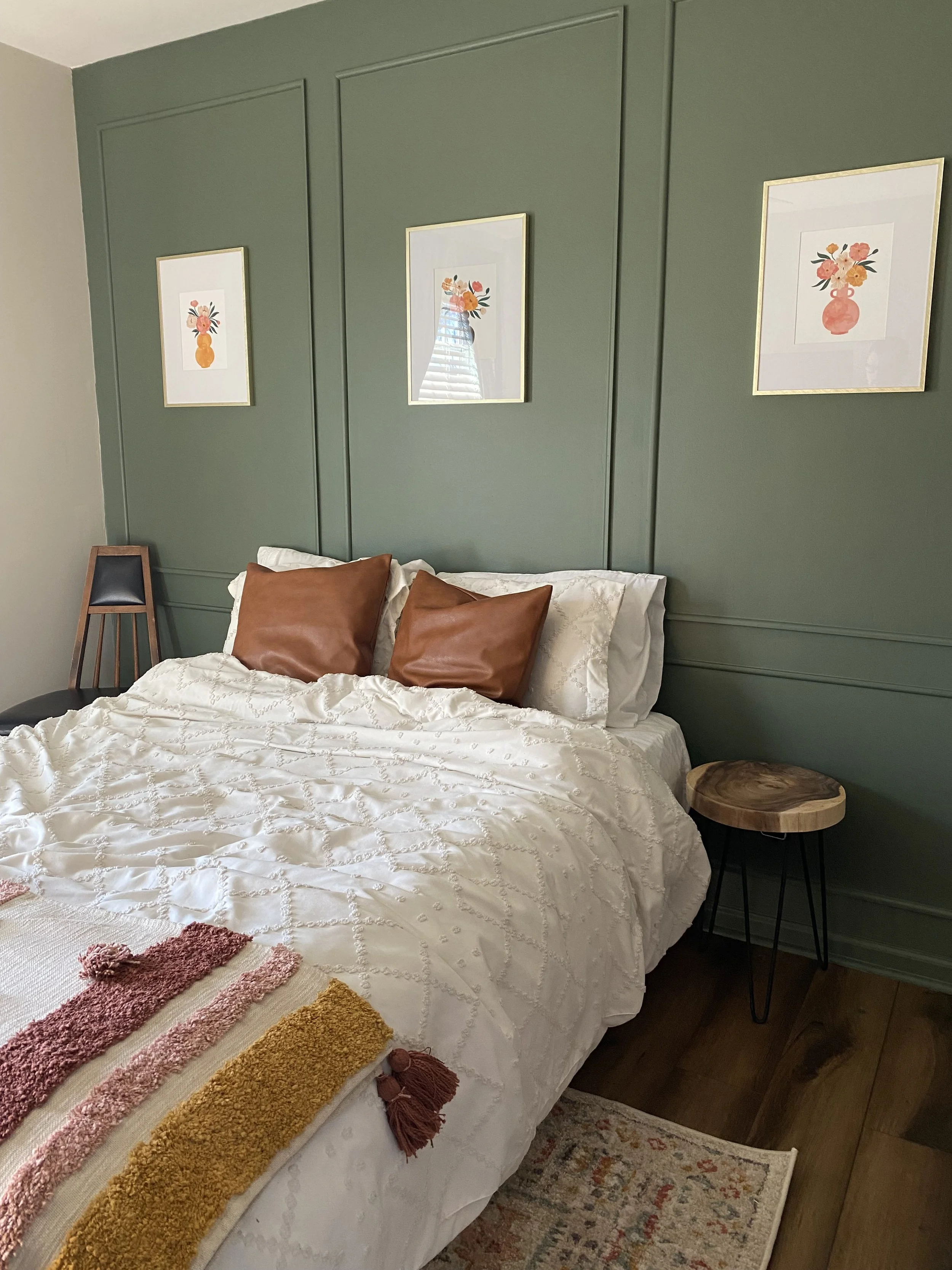 A neatly made bed with white bedding and two brown pillows, a small side table with a wooden top and black hairpin legs, an area rug in front of the bed, and three framed floral art prints above a green paneled wall.