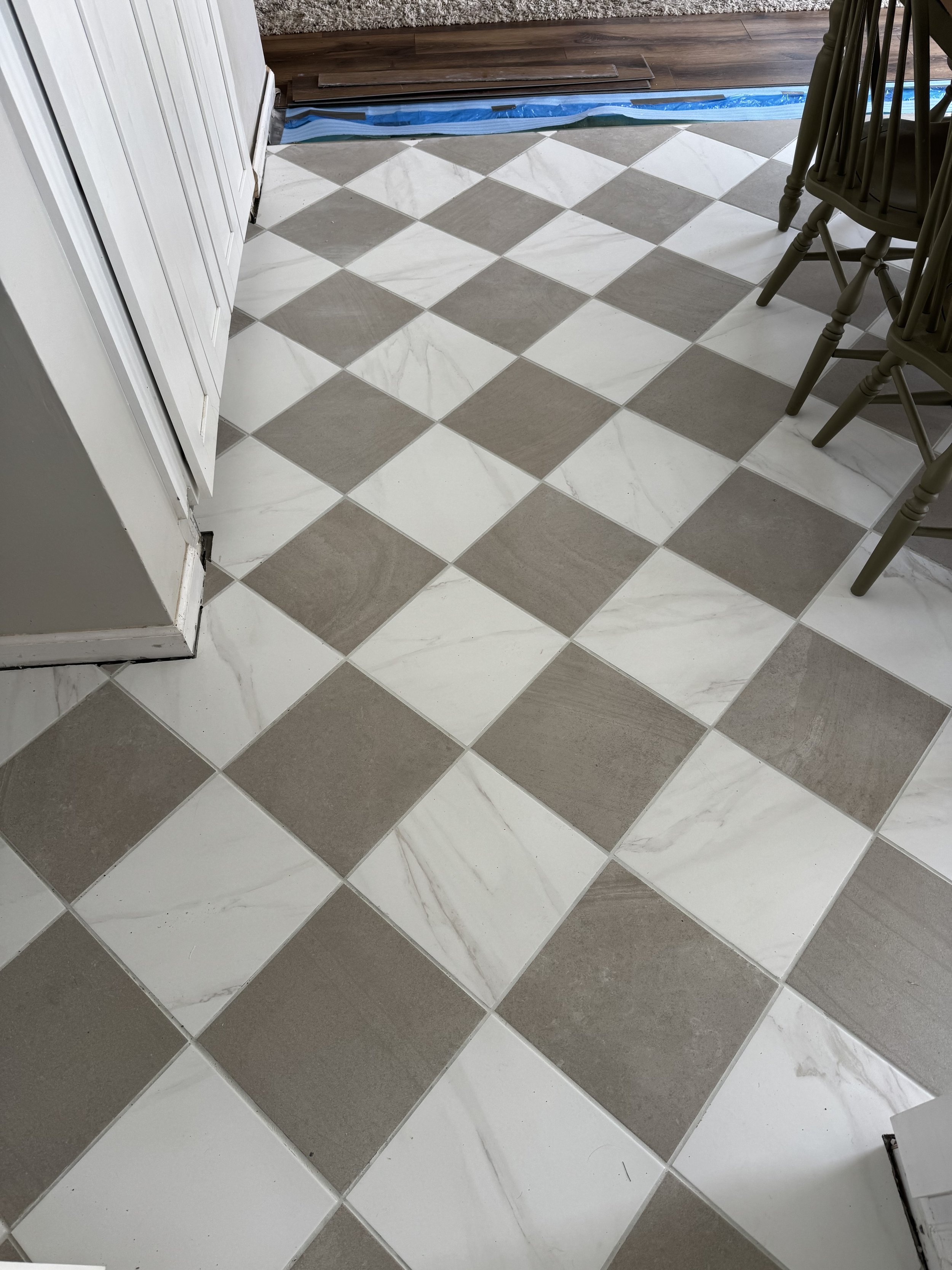 Tile floor with a checkered pattern of white and beige tiles in a room, with some wood planks and plastic sheeting visible near a wall.