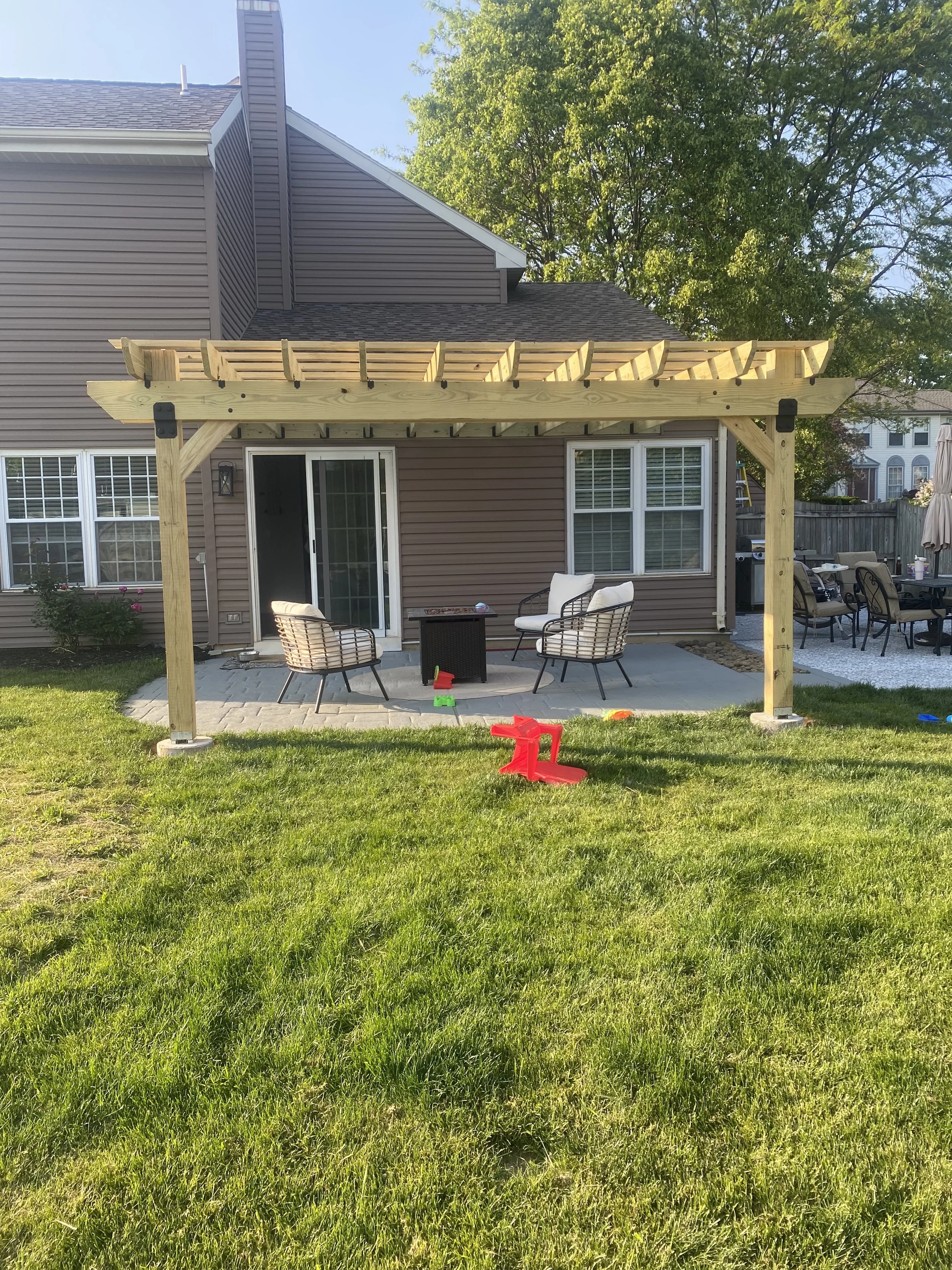 A backyard patio with a partially constructed wooden pergola. The patio has outdoor chairs and a small table. There is a grassy lawn in the foreground with a few children's toys scattered around. The house has beige siding and white-framed windows.