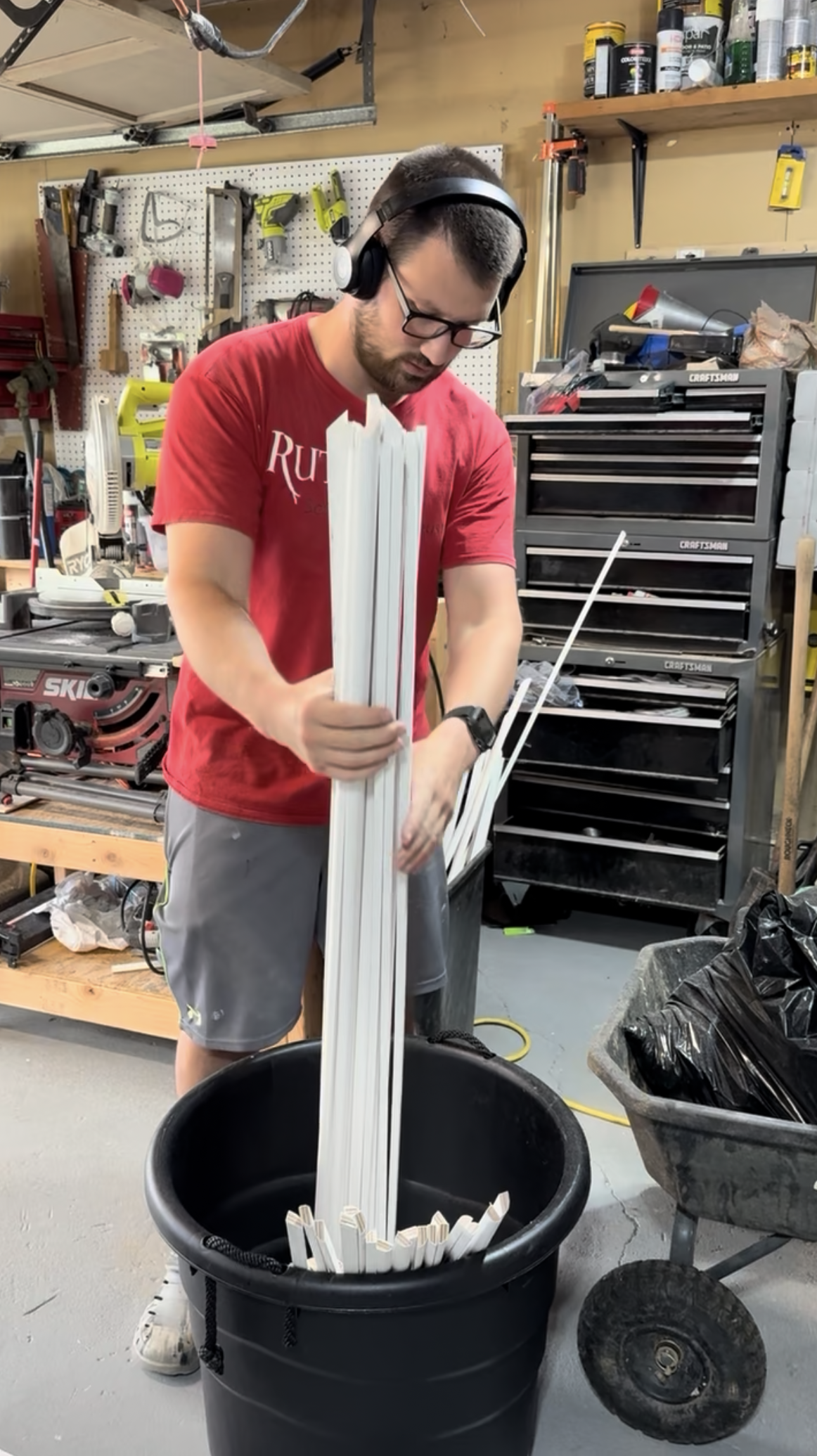 A man wearing glasses, a red t-shirt, and headphones is standing in a woodworking workshop. He is holding multiple white elongated wooden pieces above a black bucket that contains similar pieces. The workshop has tools hanging on a pegboard, a toolbo