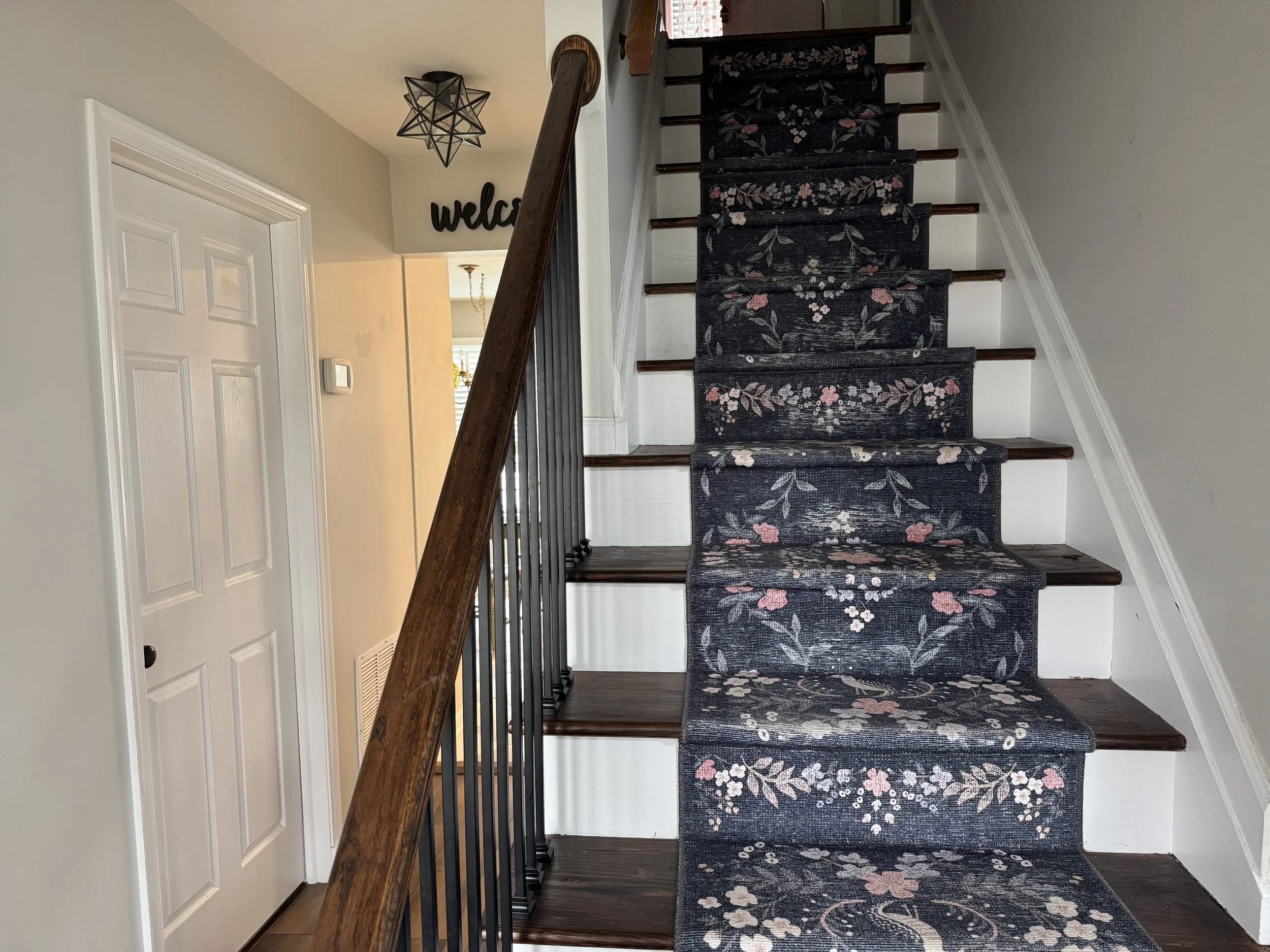 Interior view of a staircase with a dark floral carpet runner, dark wooden steps, white walls, and a wooden handrail. To the left, a white door is visible, and in the background, a decorative black 'welcome' sign hangs near a ceiling light fixture.