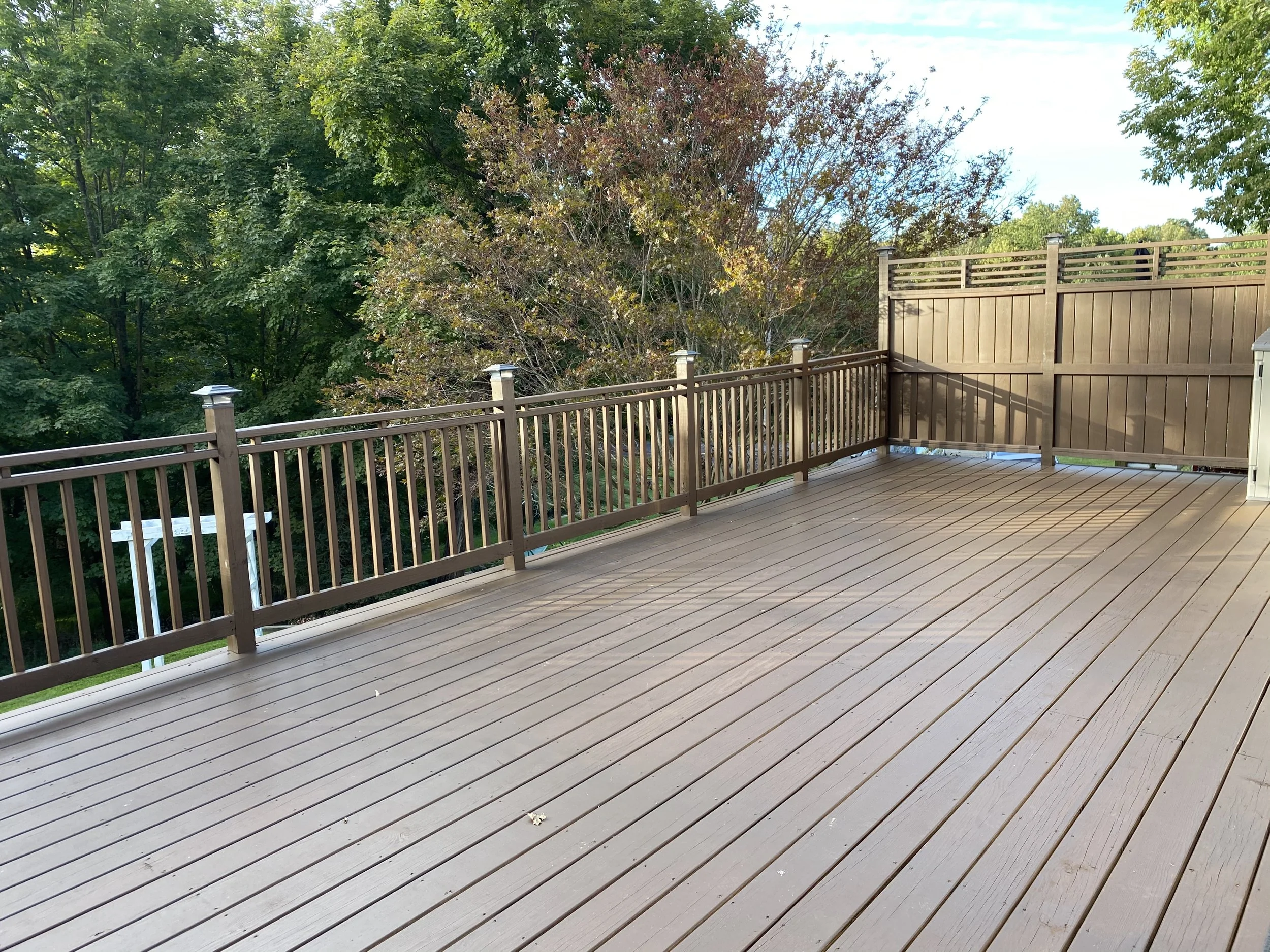 Empty wooden deck with brown railing and privacy fence, overlooking a green, leafy yard with trees.