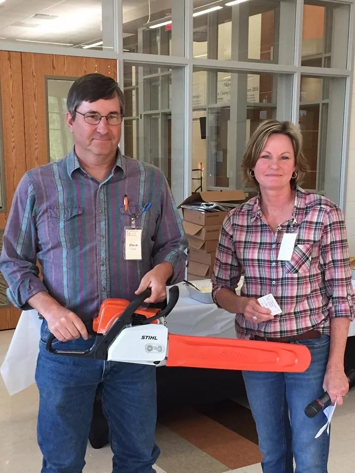 A man is holding a Stihl chainsaw he won as a door prize. They are both smiling, wearing plaid shirts, and have name tags on. There are boxes and papers on a table behind them.