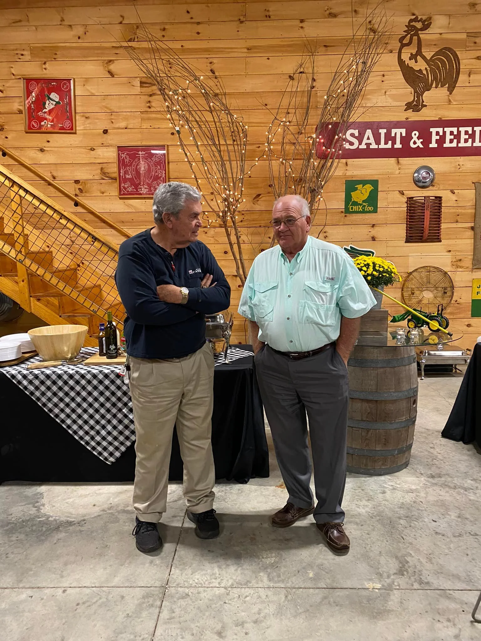 Two men standing and conversing in a rustic indoor space with wooden walls and decorations. One man has gray hair, is wearing a black jacket and khaki pants, while the other man is bald, wearing glasses, a light green shirt, and gray pants. There is a table with food and drinks, decorative branches with lights, and a wooden barrel behind them.