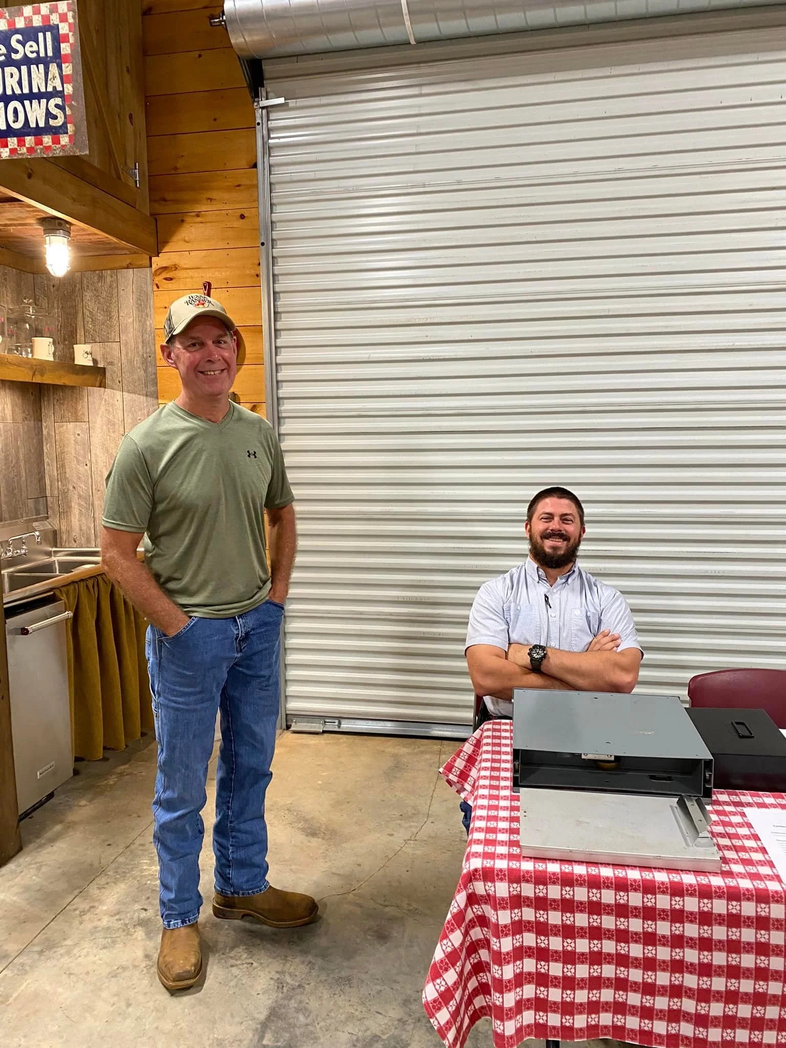 Two men in a rustic indoor setting with wood-paneled walls and a metal roll-up door. One man stands in front wearing a tan cap, green T-shirt, jeans, and boots, smiling with his hands in his pockets. The other man is seated at a table with a red-and-white checkered tablecloth, arms crossed, smiling, with a watch on his left wrist. There is a table with a printer and a tablecloth, and a small kitchen area with glasses and mugs on a shelf.