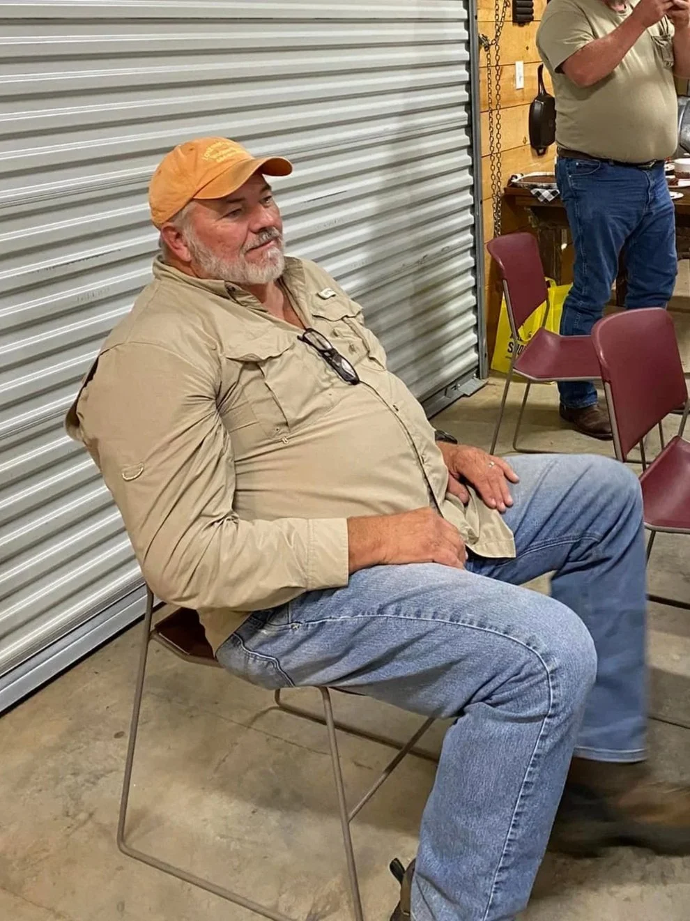 A man with a beard and glasses resting on his chest, wearing a tan cap, tan shirt, and jeans, sitting on a metal chair in a workshop or garage with a closed metal shutter backdrop.