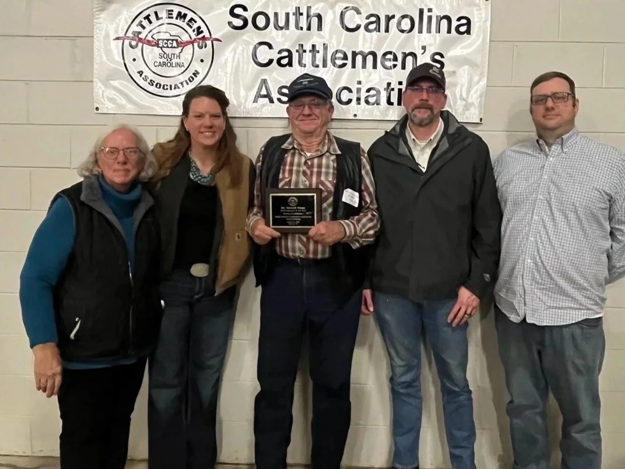 Group of five people standing in front of a South Carolina Cattlemen's Association banner, with one man in the center holding a plaque, at a conference or event.