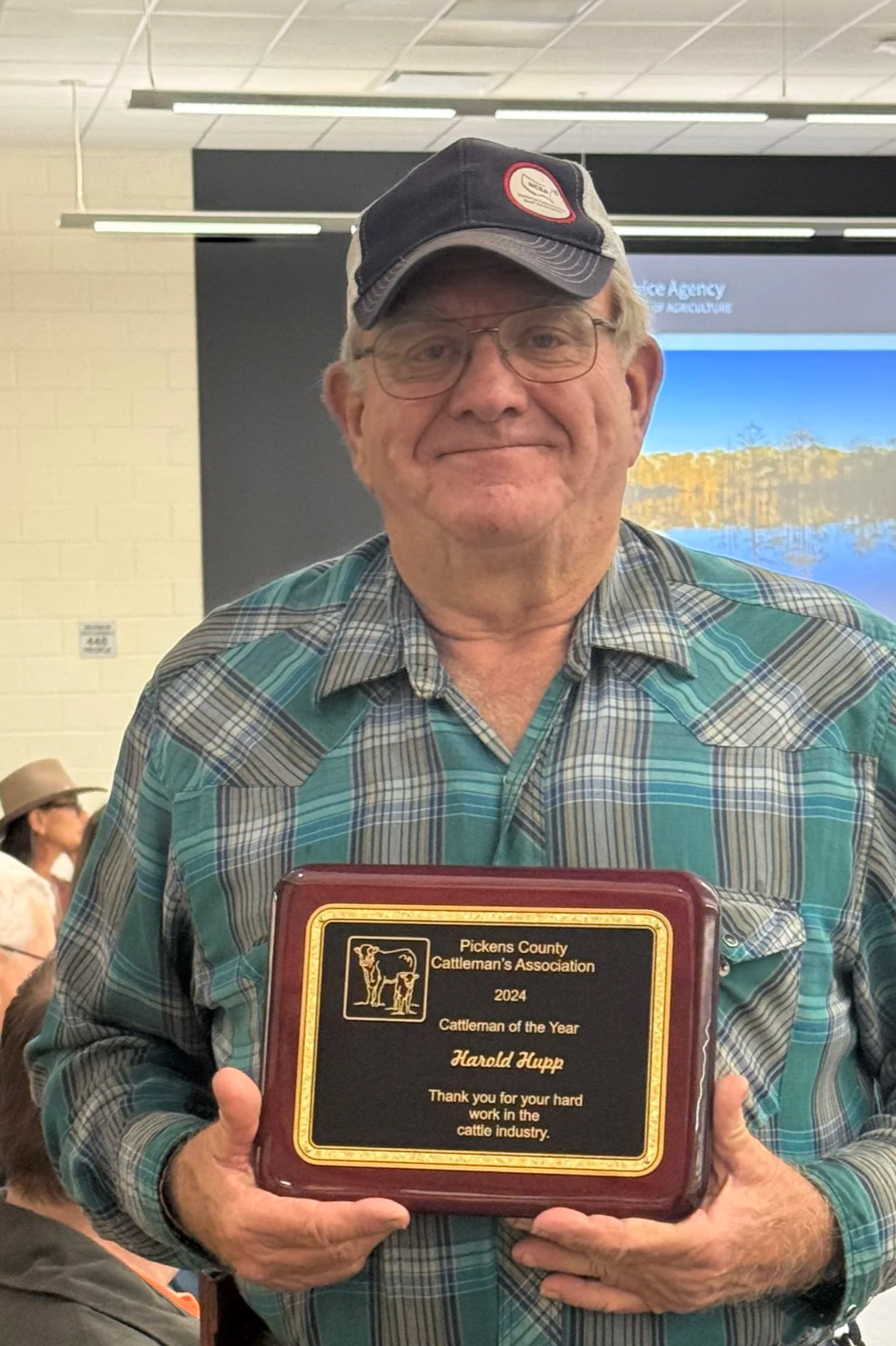 A man with glasses and a cap holding a plaque that reads 'Pickens County Cattleman's Association 2024 Cattleman of the Year Harold Hupp.' He is smiling at an indoor event with other people visible in the background.
