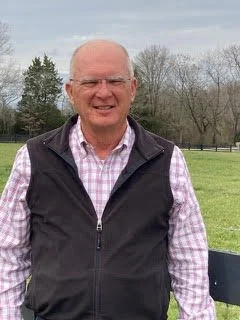 An elderly man standing outdoors in a park, smiling, wearing glasses, a purple checkered shirt, and a black vest.