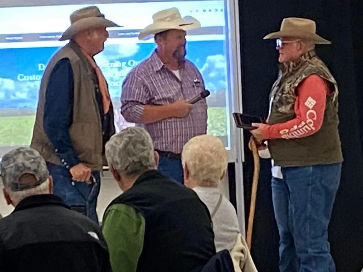 Three men wearing cowboy hats and outdoor clothing are standing and talking to each other in front of an audience, with a presentation screen behind them. One man holds a microphone, and the other has a tablet. Several audience members are seated, wearing jackets and hats.