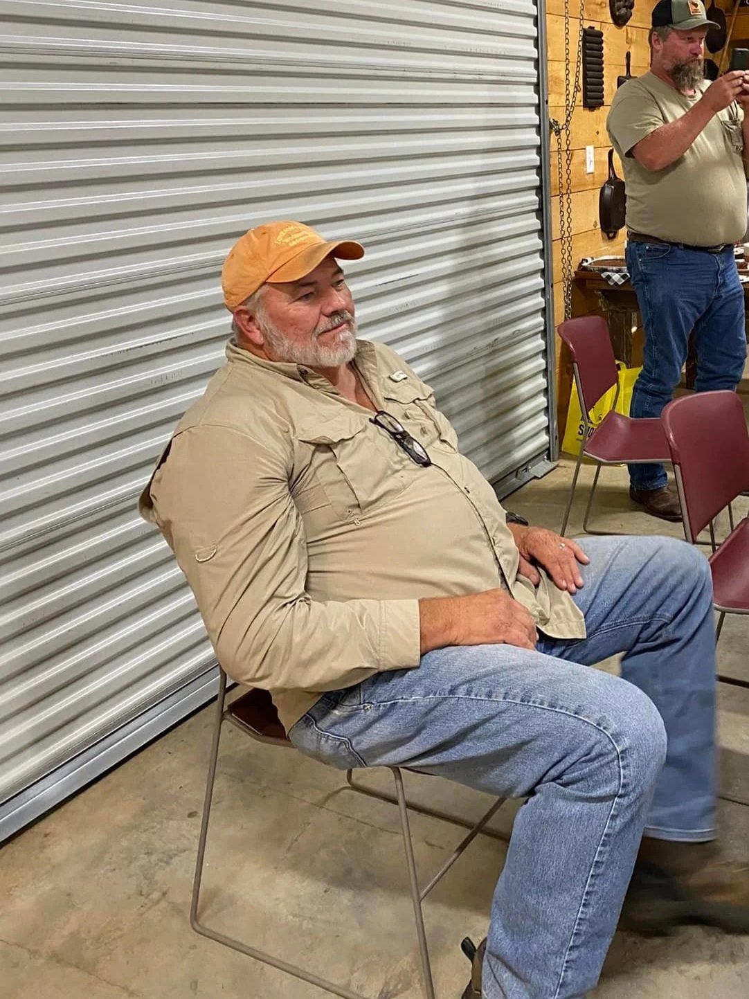 A middle-aged man with a beard and mustache sitting on a metal chair, wearing a tan jacket, jeans, and an orange cap, in a room with a metal garage door and wooden walls. Another man in a baseball cap and t-shirt is standing nearby, taking a photo.