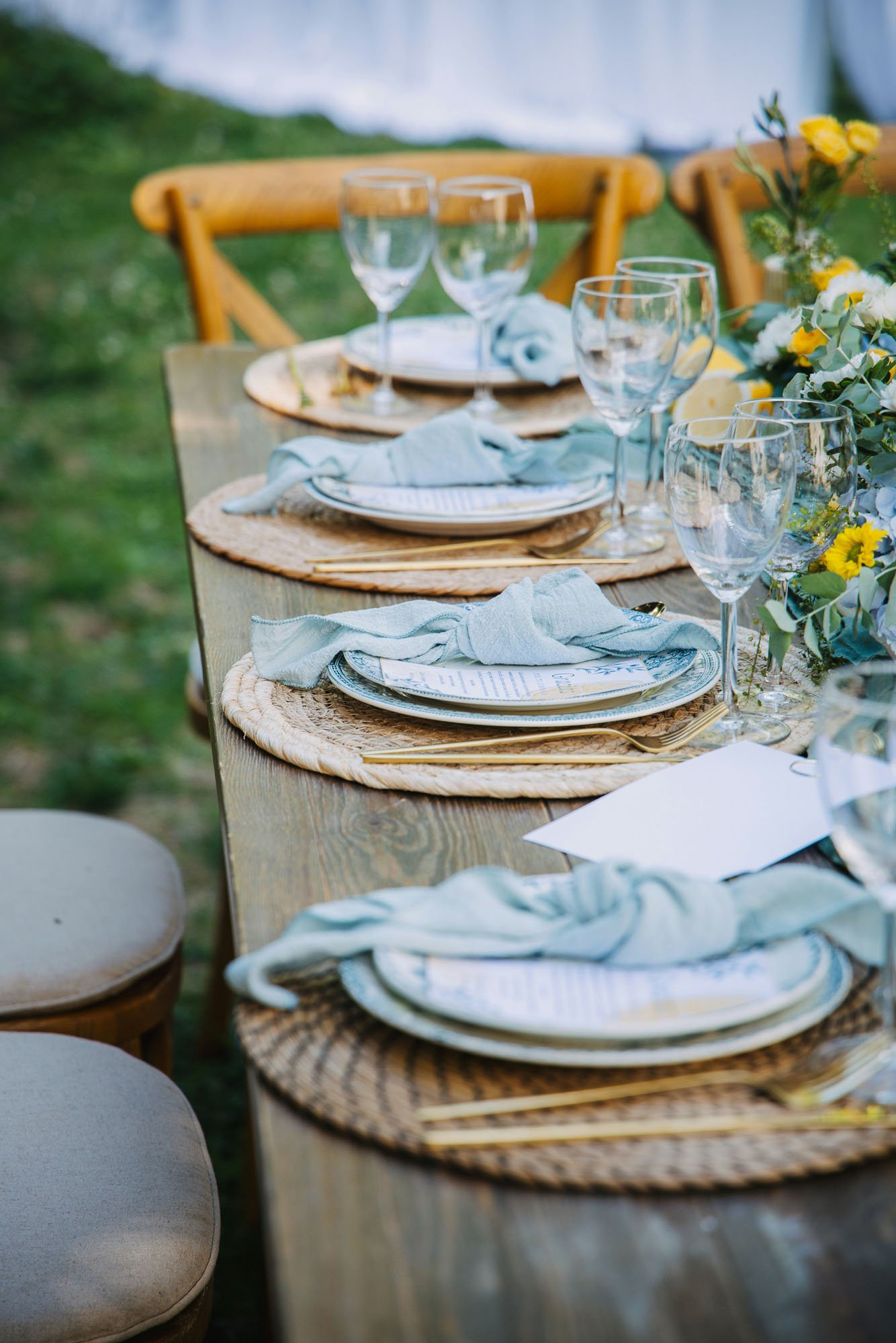 A rustic outdoor dining table set with woven placemats, white plates with printed menus and light blue napkins, wine glasses, cutlery, and a floral centerpiece with yellow and white flowers.
