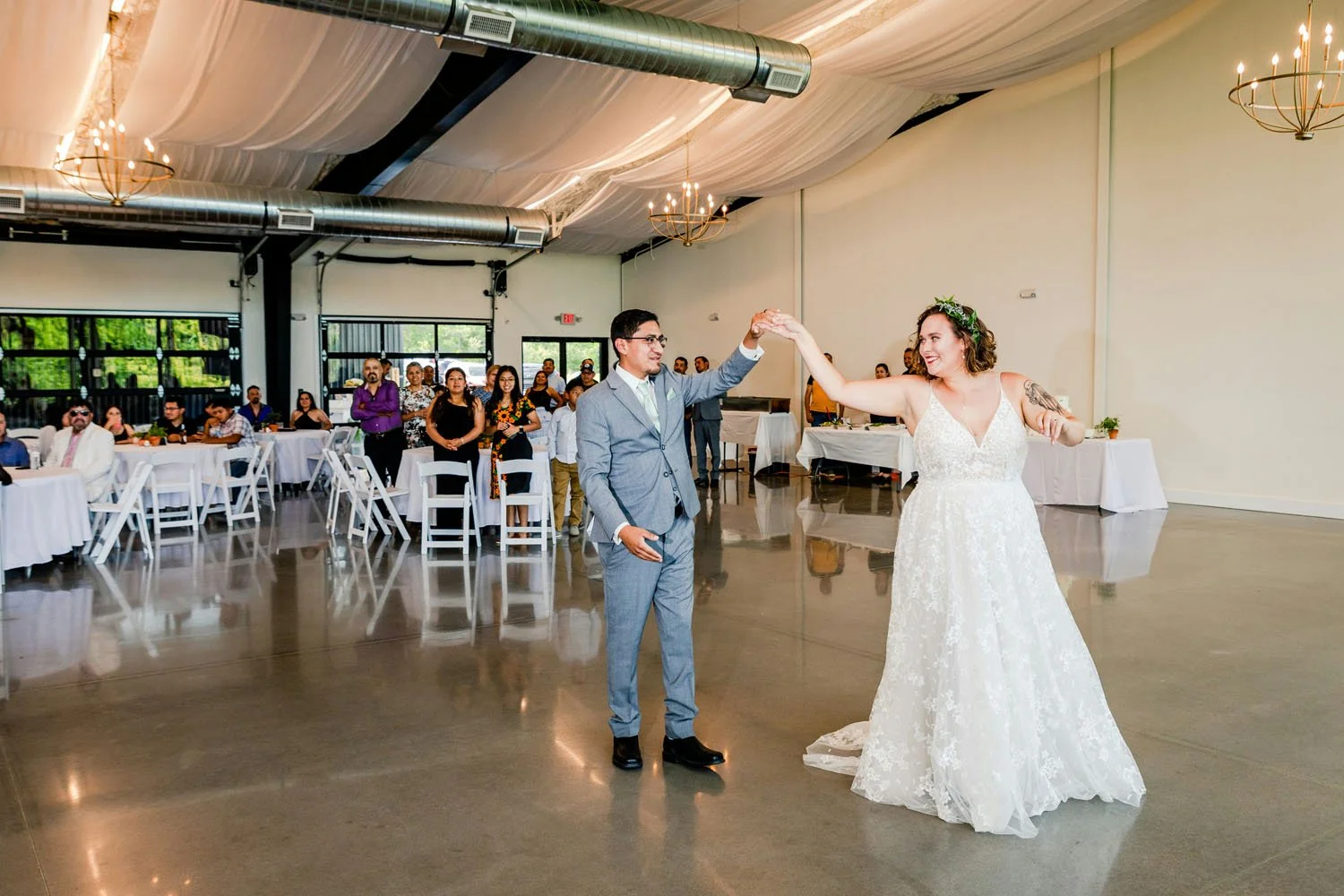 A bride and groom are dancing during their wedding reception in a large hall. The bride is wearing a white wedding gown with lace, and the groom is in a light gray suit. Guests are seated at round tables and standing in the background, watching the couple dance.
