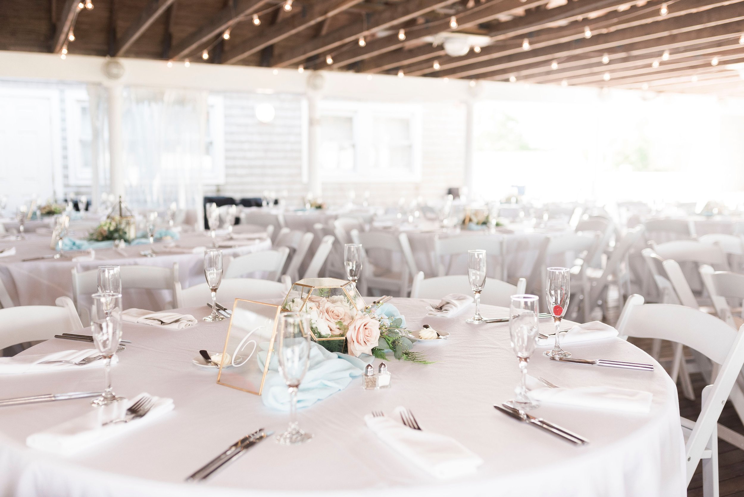 Wedding reception hall with round tables covered in white tablecloths, set with glassware, silverware, and folded napkins, decorated with floral centerpieces and framed photos, illuminated by natural light and string lights.
