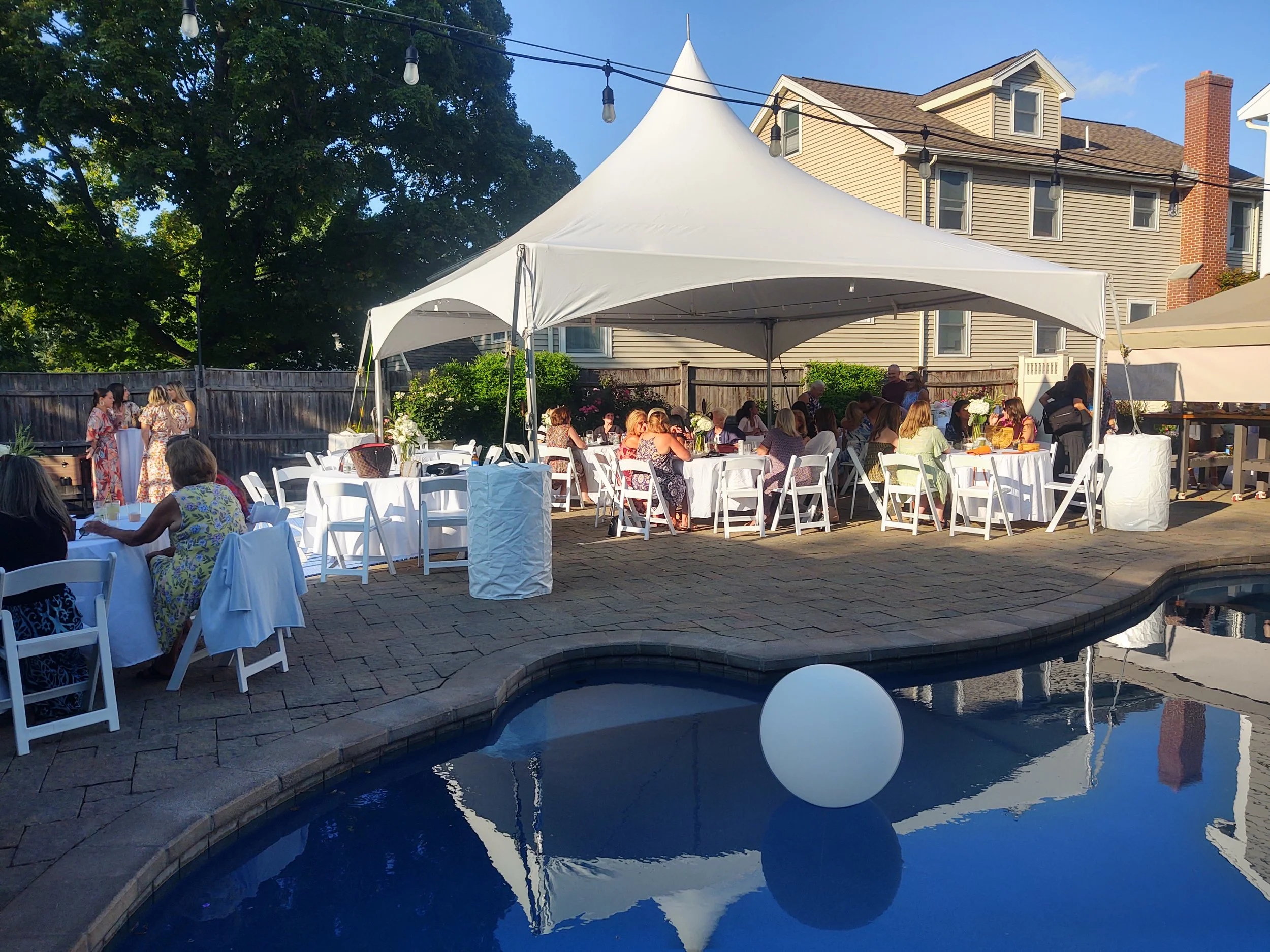A backyard party with white tables and chairs, a large white canopy tent, string lights, and a swimming pool in the foreground, with a house and trees in the background.