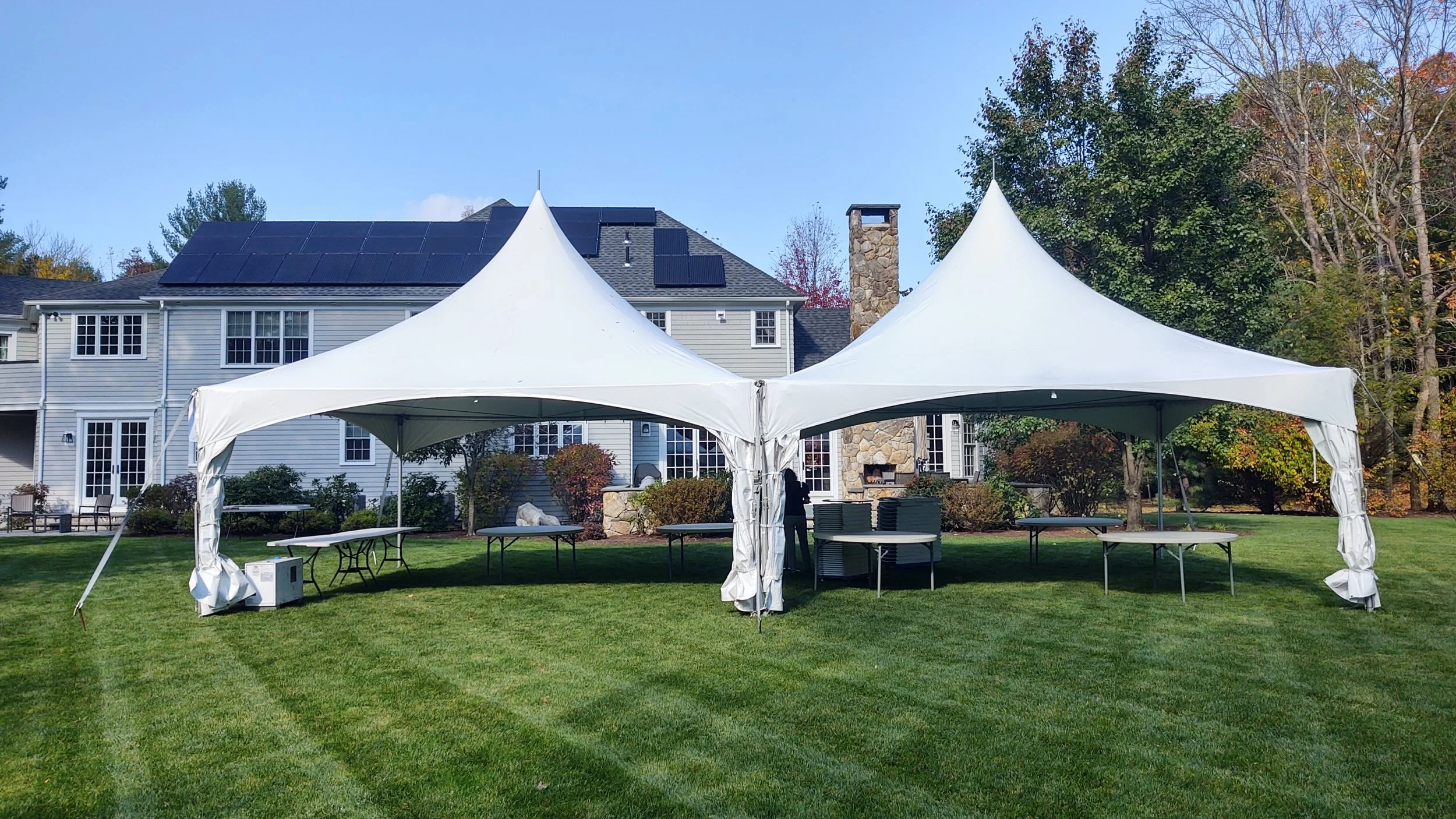 A large white event tent set up on a well-manicured lawn in front of a house, surrounded by trees with fall foliage.