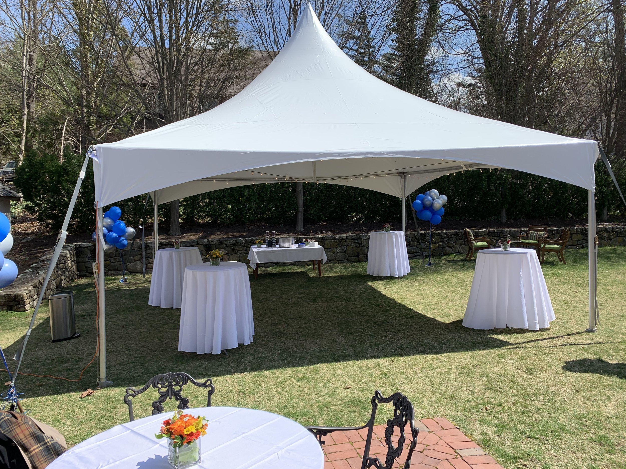 Outdoor event setup with a large white tent, high-top tables with white tablecloths, balloons, and chairs on a grassy lawn surrounded by trees.