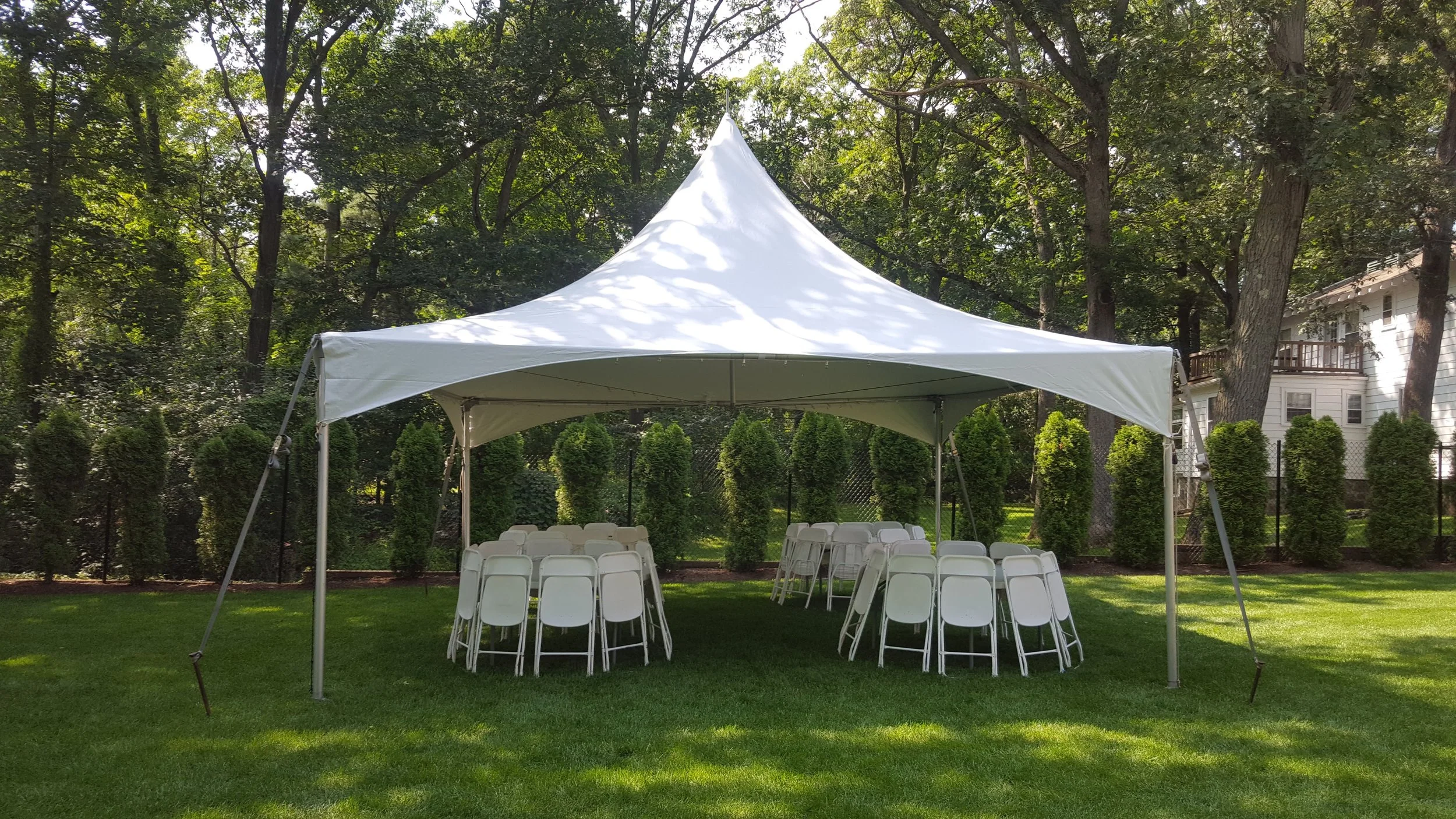 White outdoor event tent set up on a grassy lawn with foldable chairs arranged underneath, surrounded by trees and a residential house in the background.
