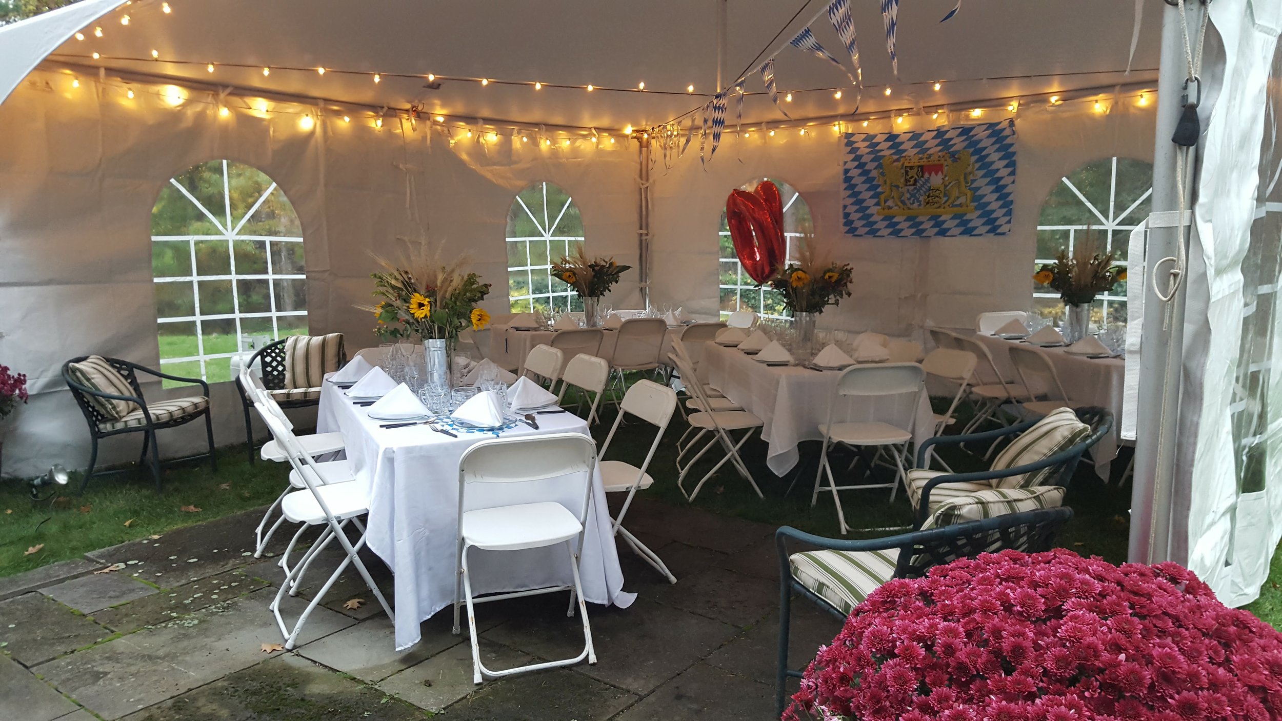 Decorated outdoor tent with long dining tables, white tablecloths, flower centerpieces, and chairs, featuring string lights.
