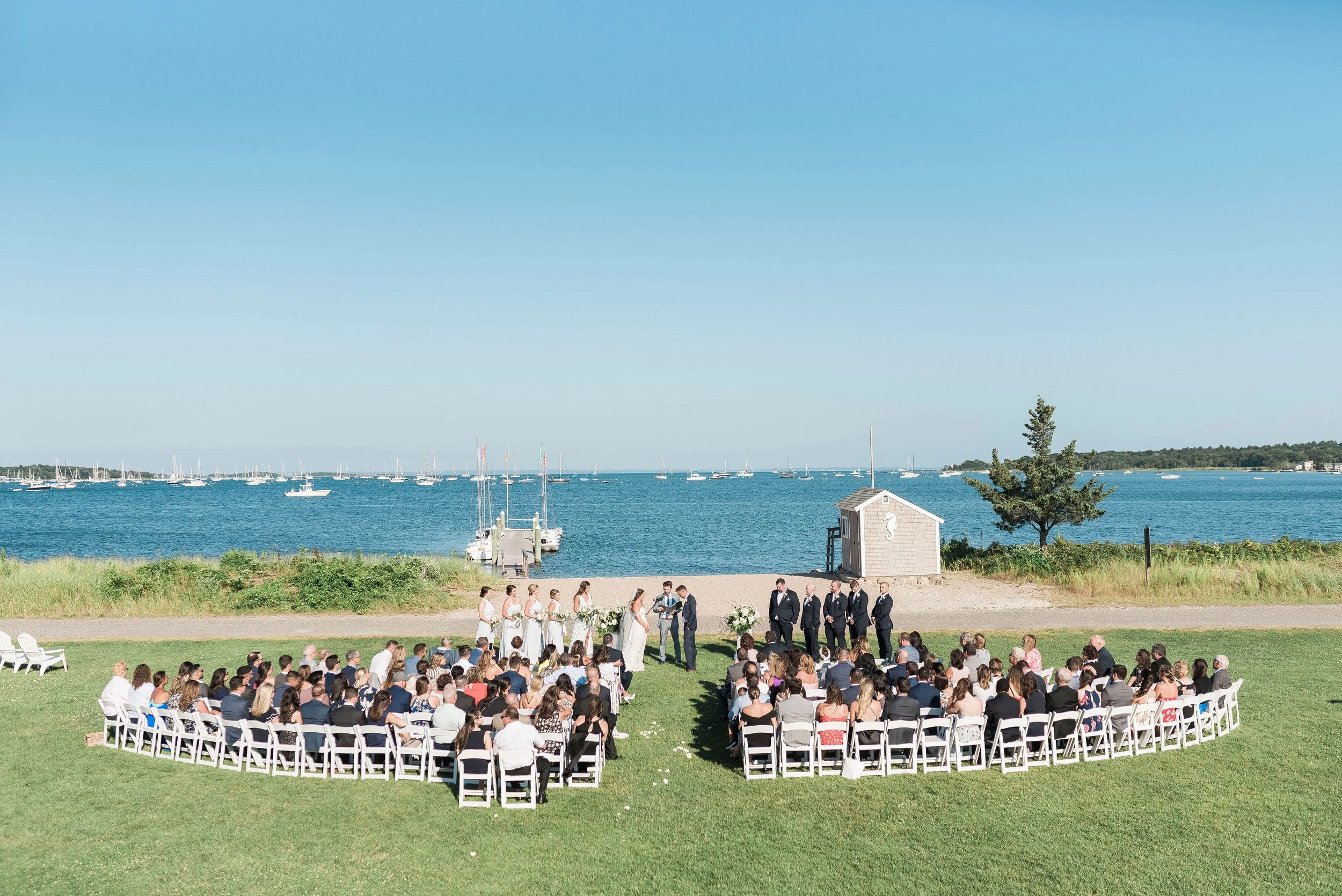 Outdoor wedding ceremony on a grassy area near the water, with guests seated in white chairs, a bride and groom exchanging vows, and a backdrop of a lake with sailboats and a small dock.