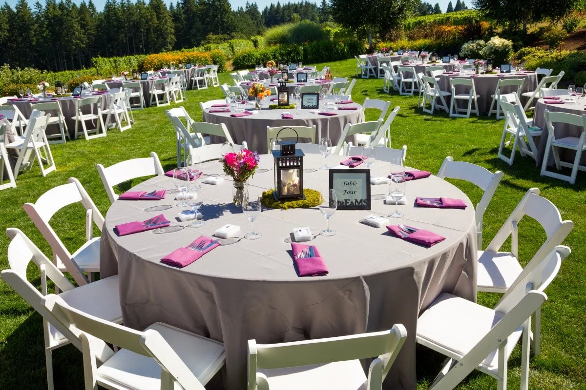 Outdoor wedding reception setup with round tables decorated with flowers, lanterns, pink napkins, and all white chairs on a lush green lawn with a background of trees and bushes.