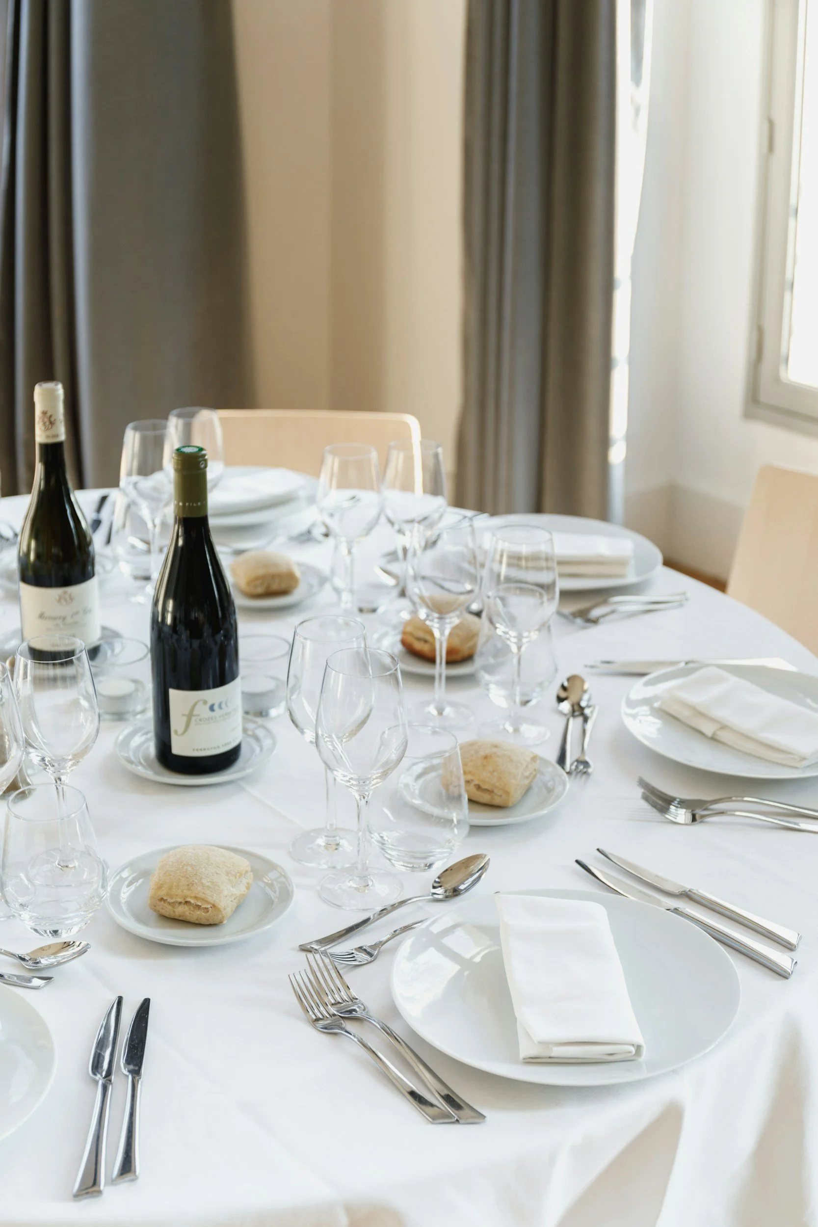 A formal dining table set with white tablecloth, multiple wine glasses, two wine bottles, plates with bread rolls, and silverware, in a well-lit room with curtains and windows.