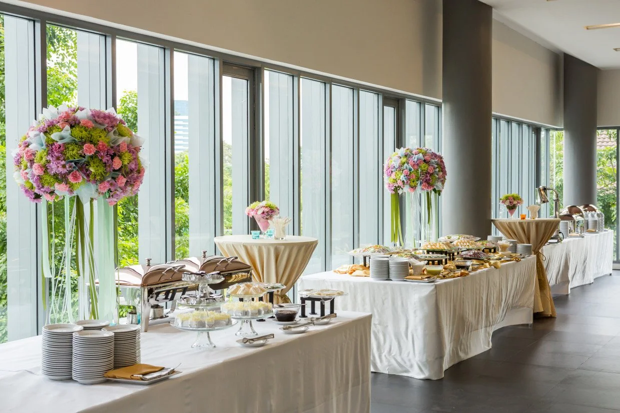 Buffet table set up with floral centerpieces, plates, and food options in a bright, modern room with large windows and greenery outside.