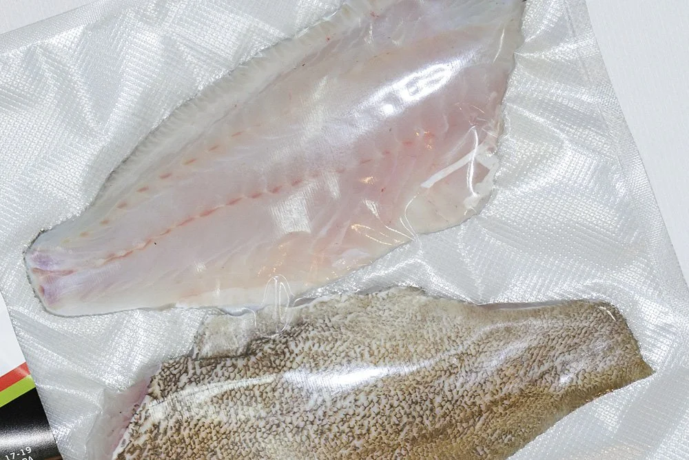Two vacuum-sealed fish fillets, one with light-colored flesh and the other with darker, patterned skin, on a textured surface.