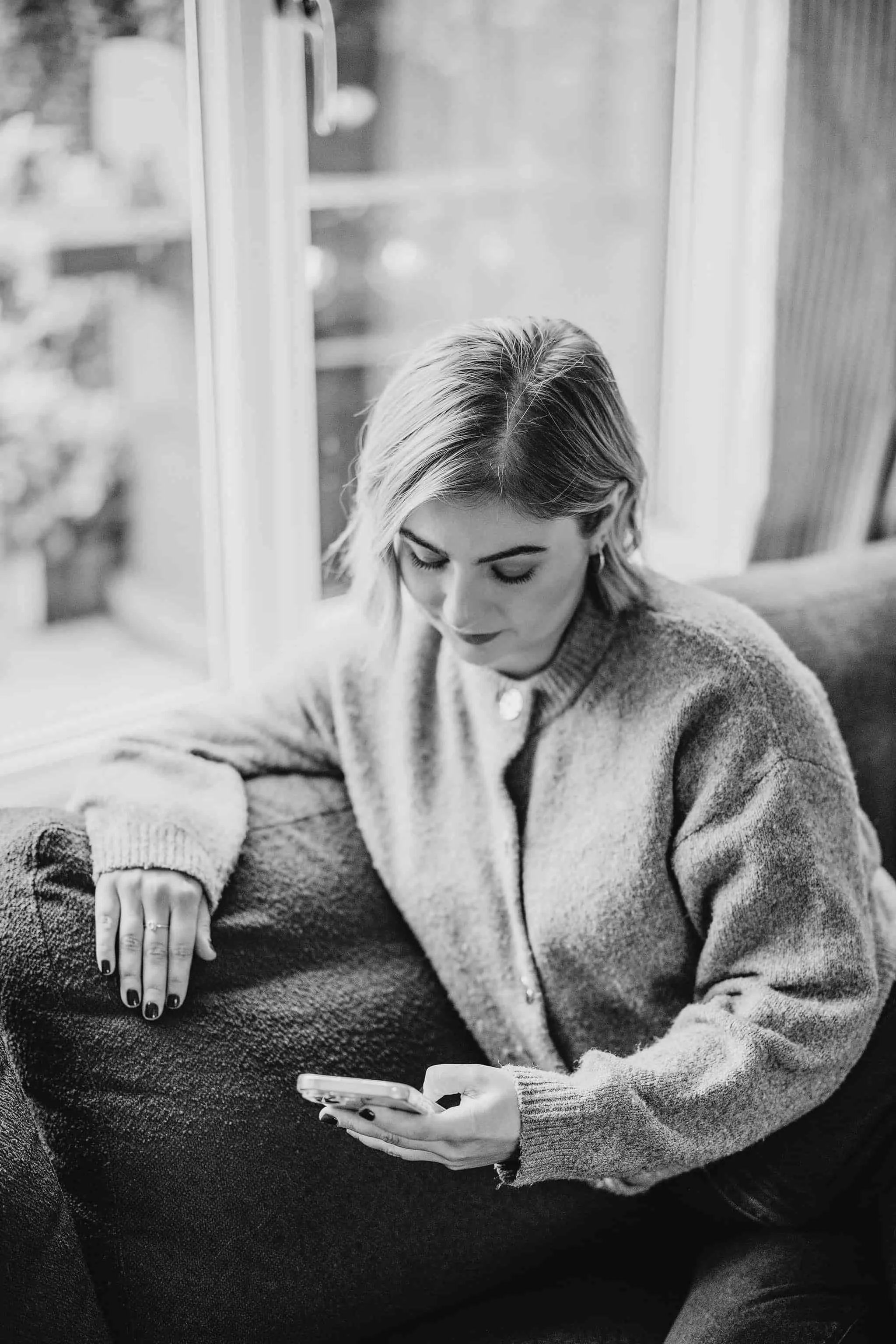 A woman with short blonde hair, wearing a sweater, sitting on a couch near a window, looking at her phone.