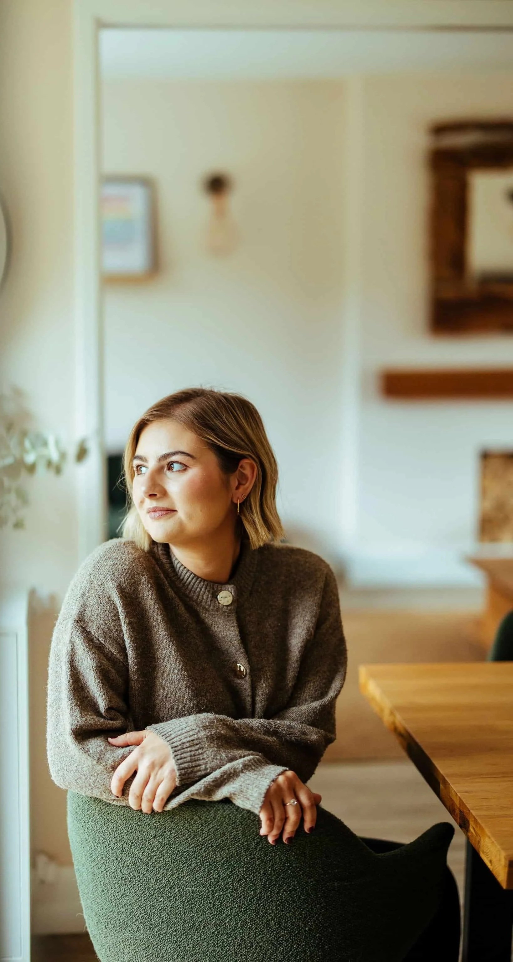 A woman with shoulder-length hair wearing a brown sweater sitting at a wooden table in a cozy indoor setting.