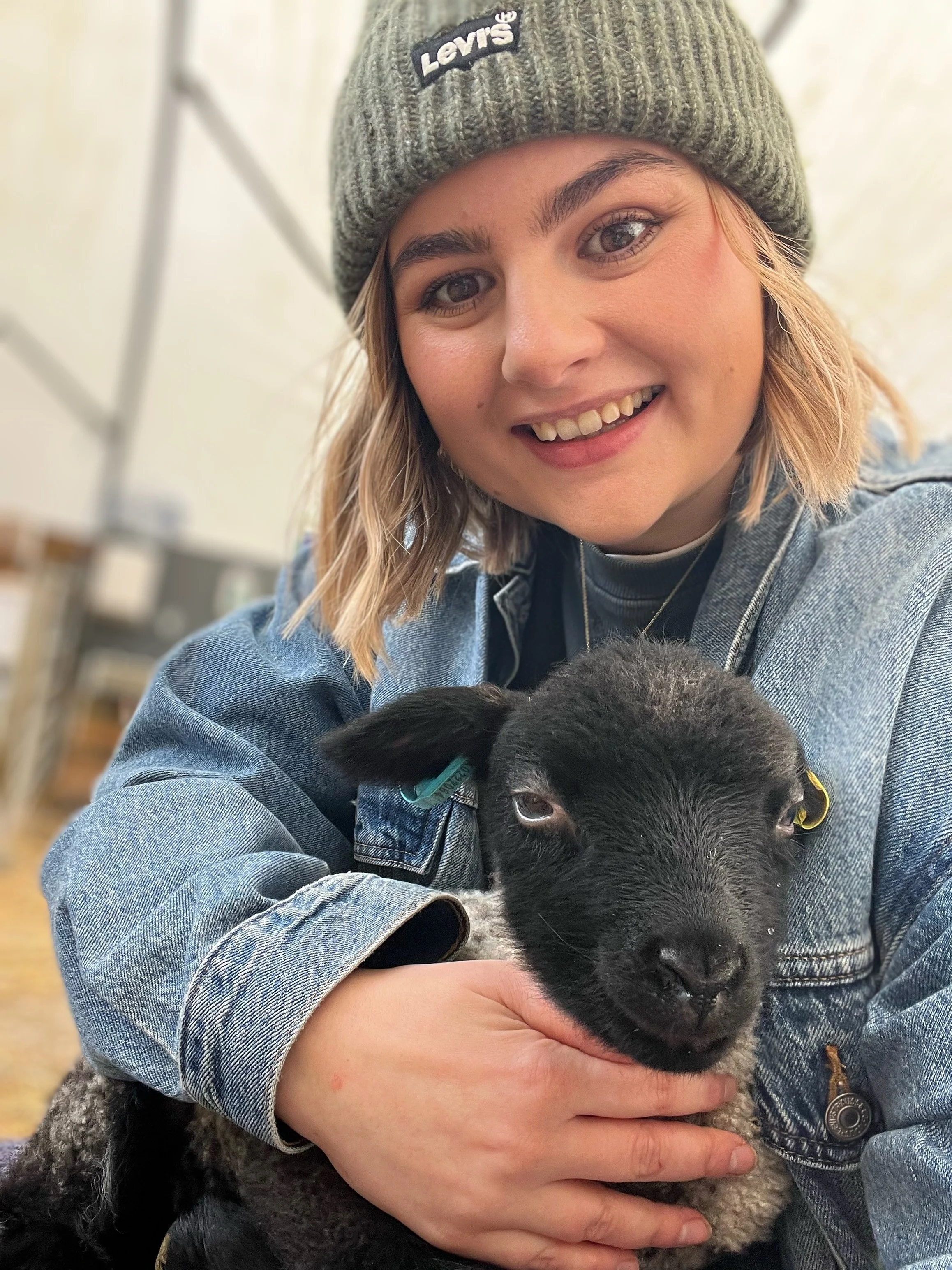 A young woman in a gray beanie and denim jacket holding a black puppy with floppy ears and gray fur on the chest, smiling at the camera.