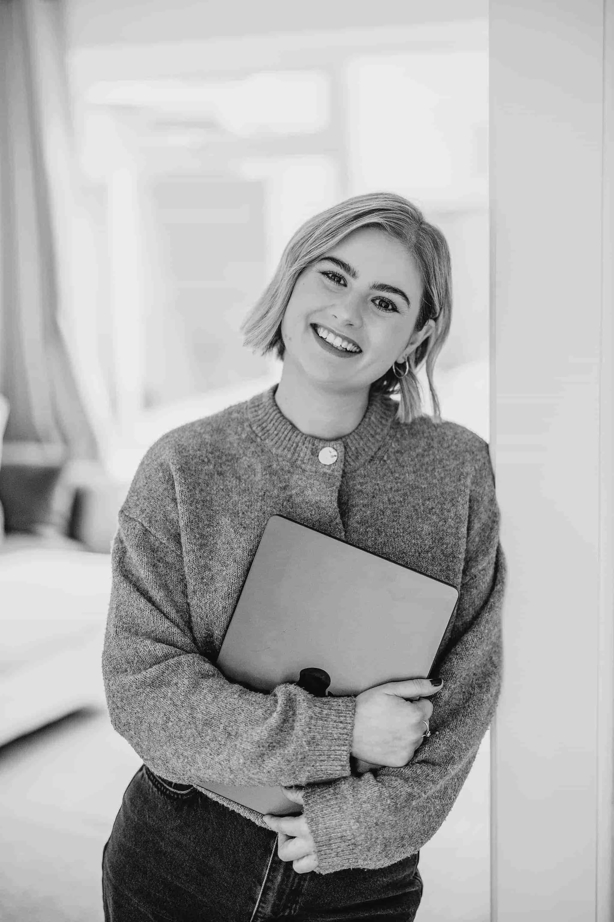 Black and white photo of a smiling woman with short hair, holding a laptop, standing near a window with curtains in a cozy room.