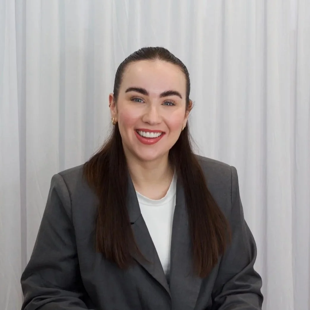 Smiling woman on a neutral curtain backdrop, wearing a grey suit jacket.